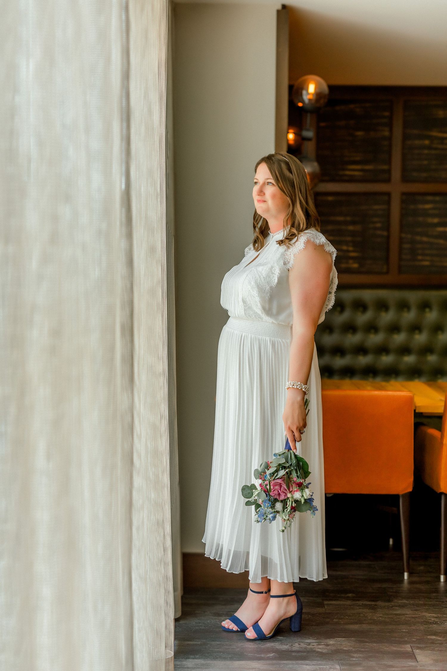 Bride in white midi dress holding floral bouquet standing by window in elegant venue wearing blue heeled sandals.