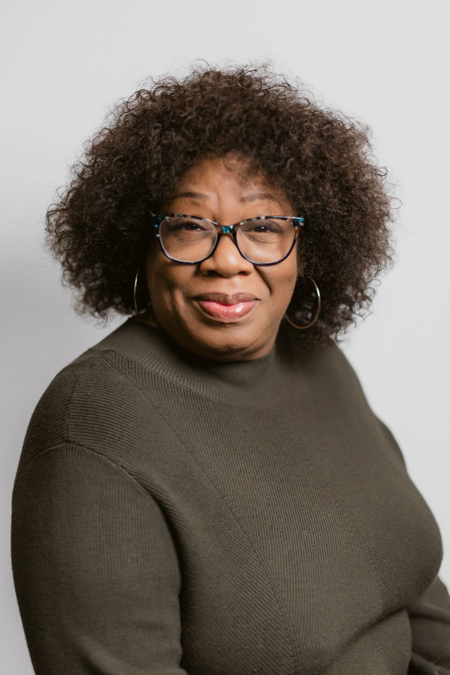 Professional headshot of person in olive turtleneck sweater and glasses against grey background.