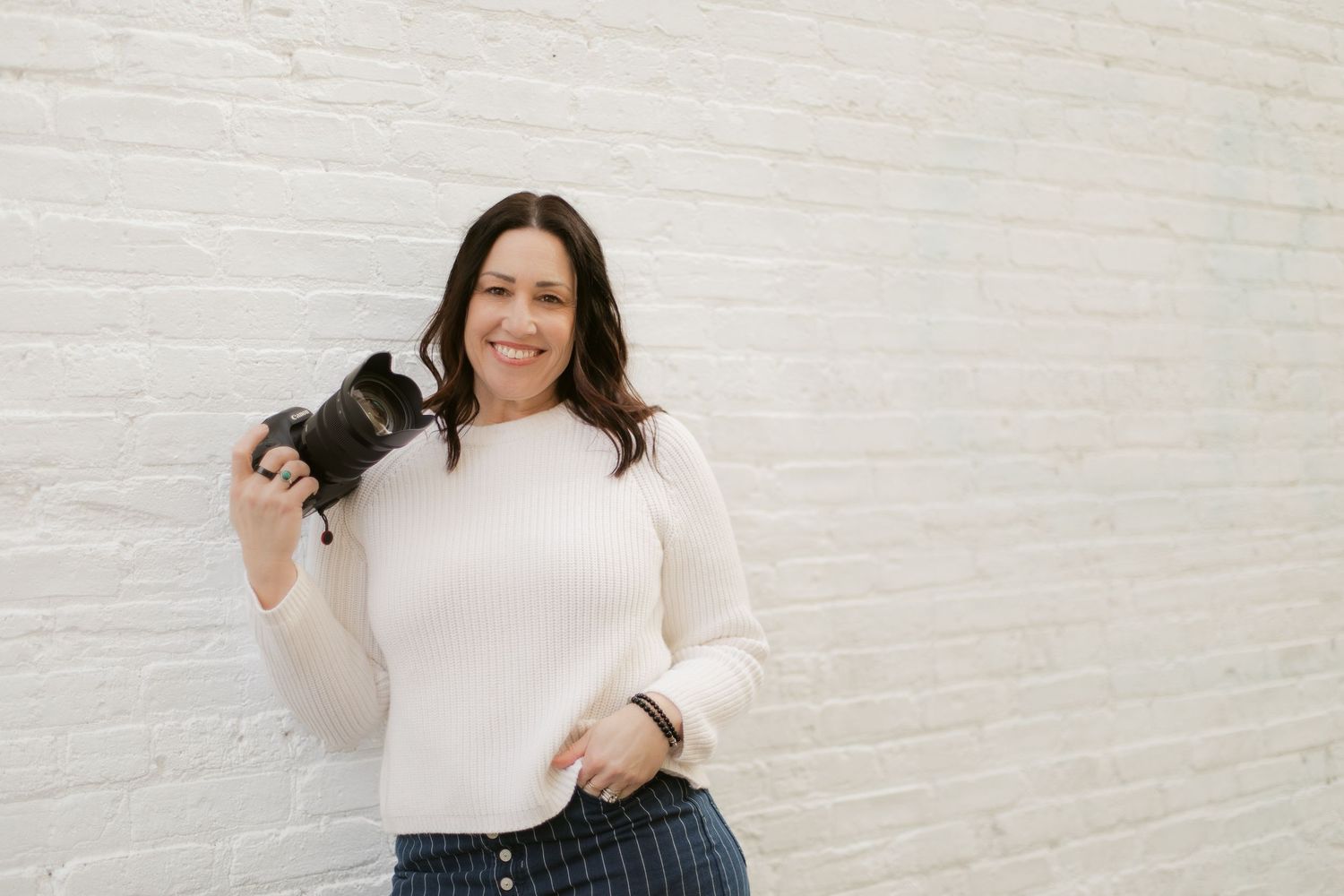 Smiling woman in white sweater holding a camera against a white brick wall.