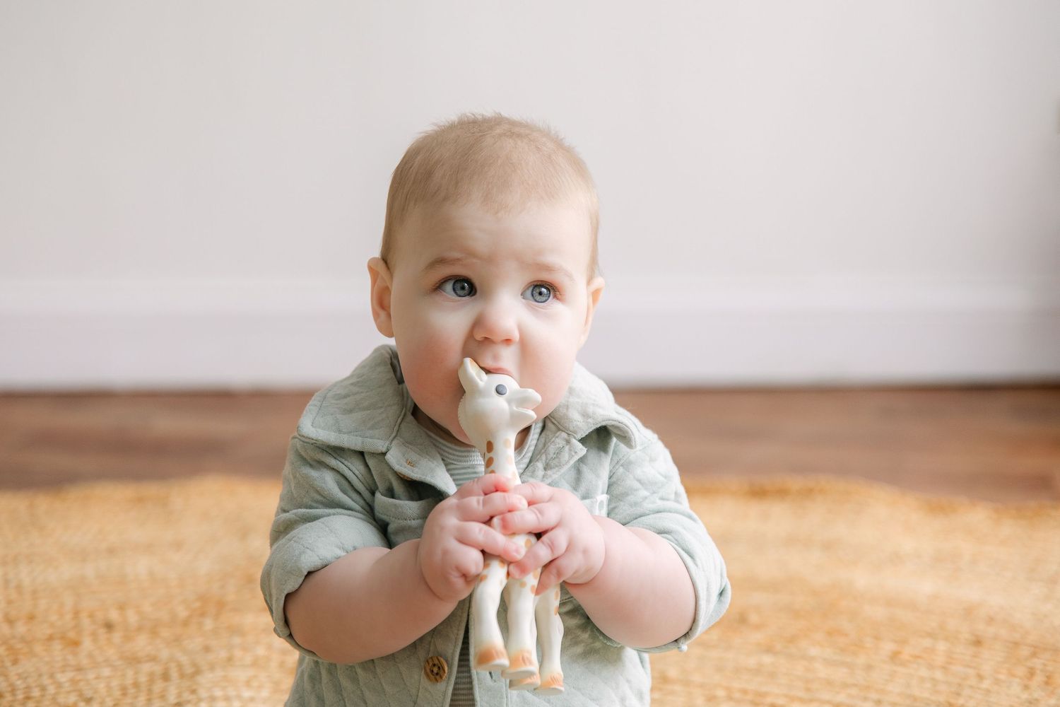 A baby in a sage green outfit holds and drinks from a bottle while sitting on a beige carpet.