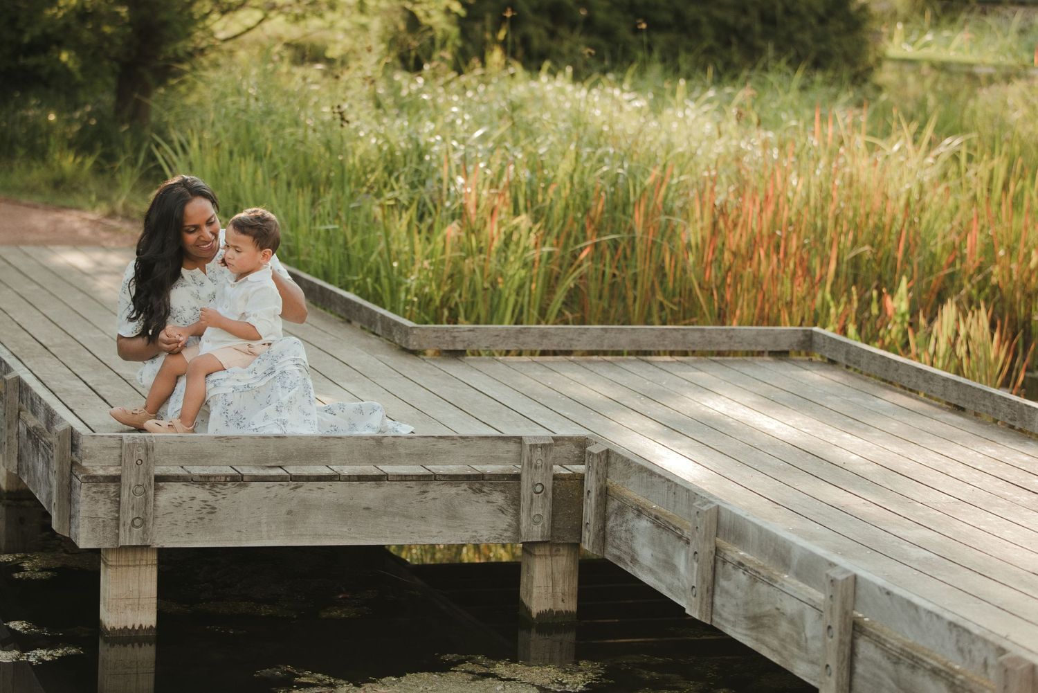 Mother and daughter sitting together on a wooden dock surrounded by lush greenery and tall reeds near a calm pond.