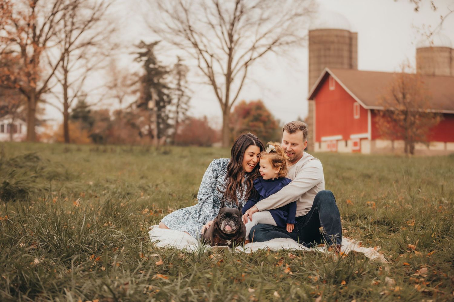 A couple sits with their dog on a blanket in front of a red barn on an autumn afternoon.