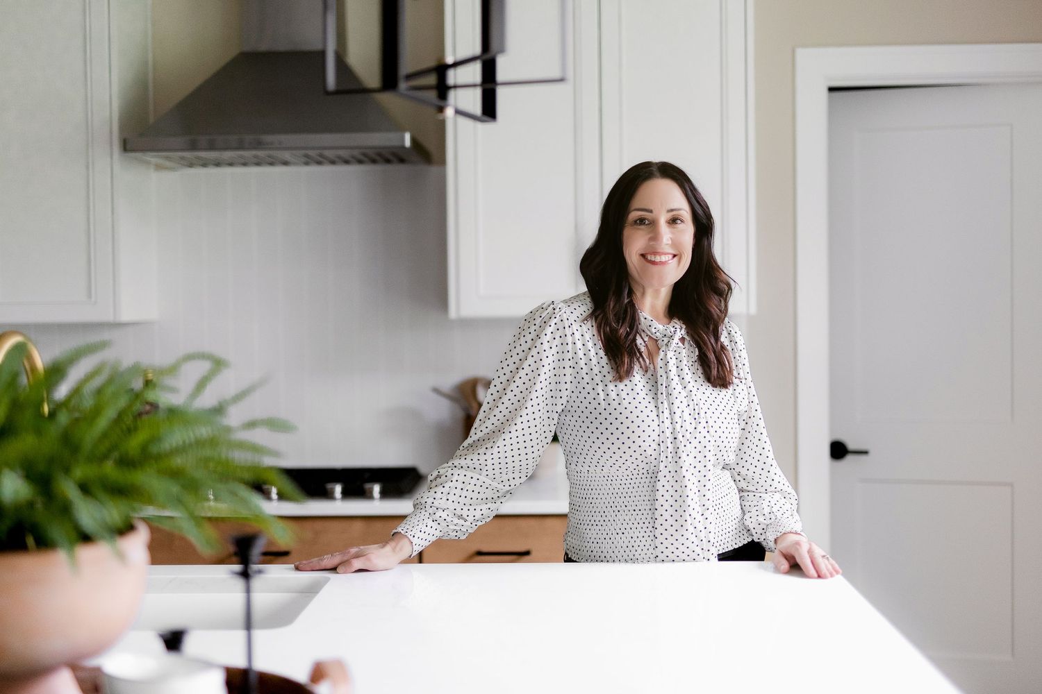Professional in polka dot blouse stands at white counter in modern kitchen setting.