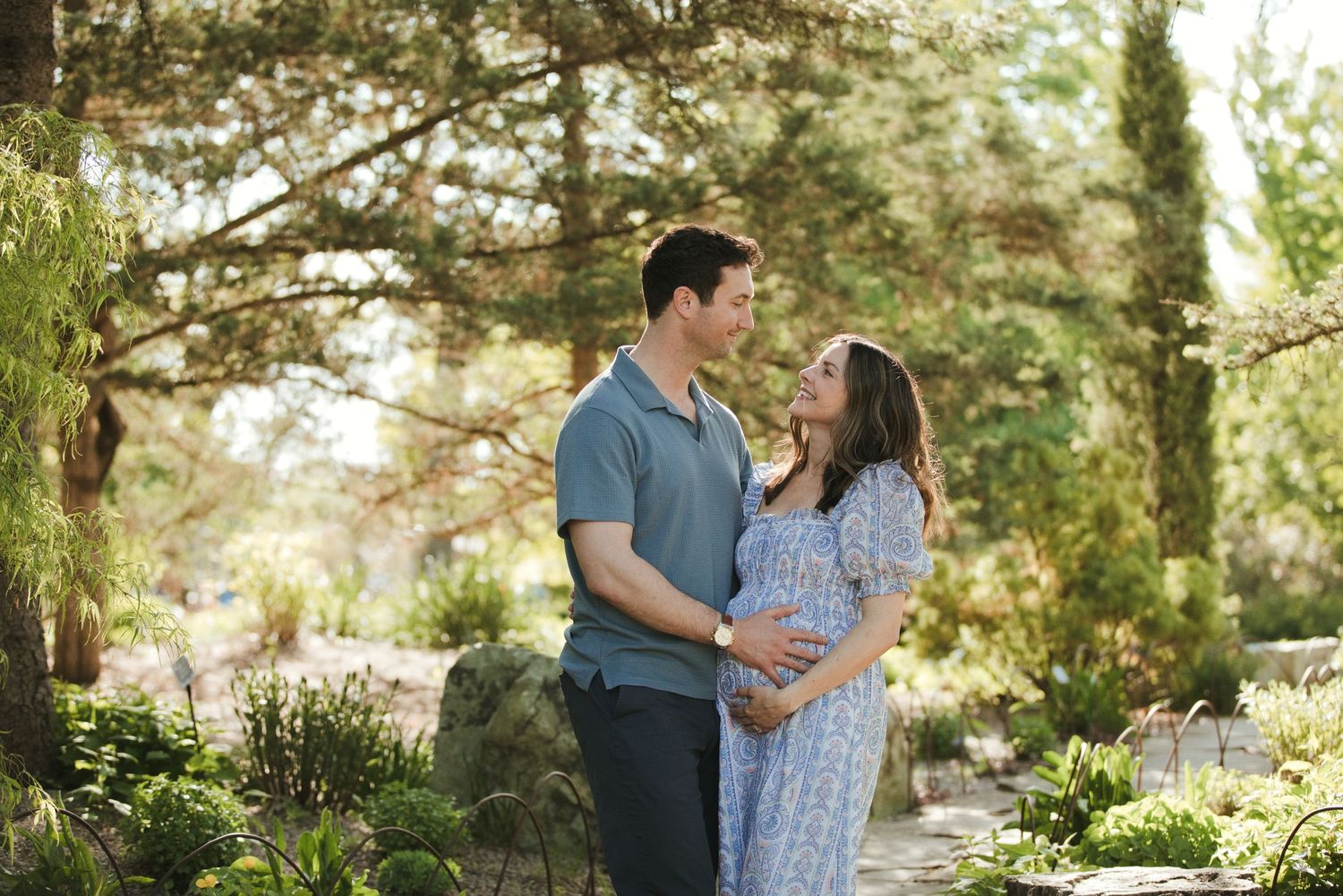 Couple sharing a tender moment in a lush garden, man holding pregnant woman's belly in soft natural light.