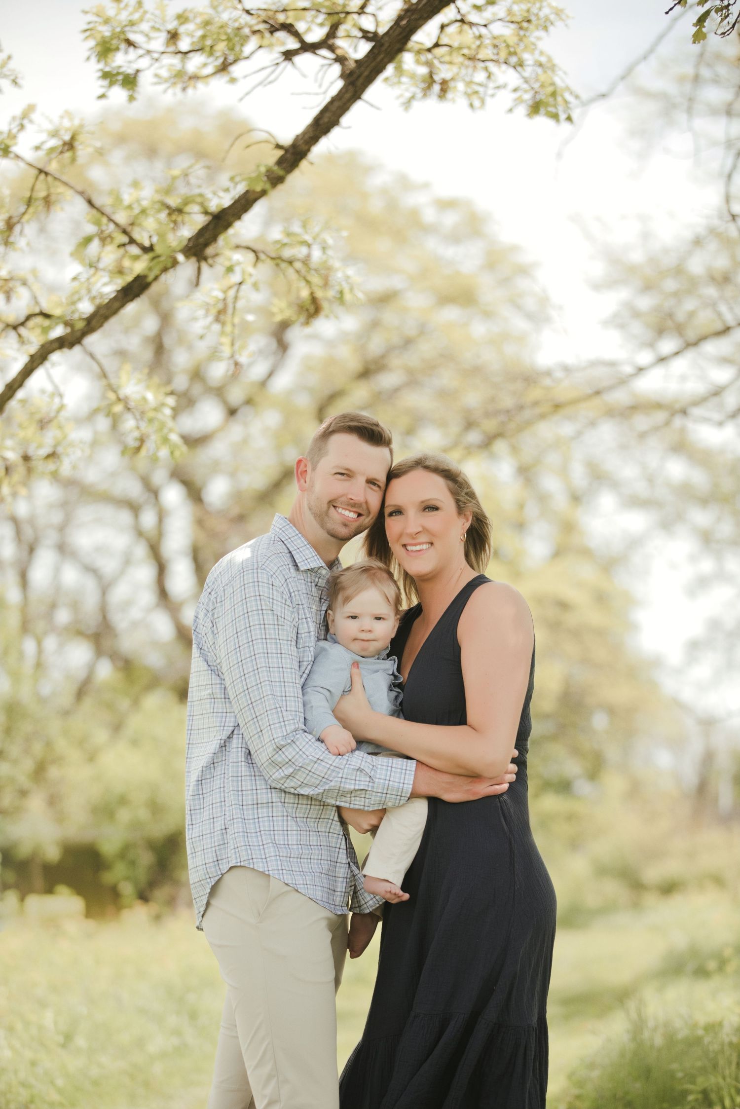A smiling couple holds their baby outdoors under blooming trees in a bright spring family portrait session.