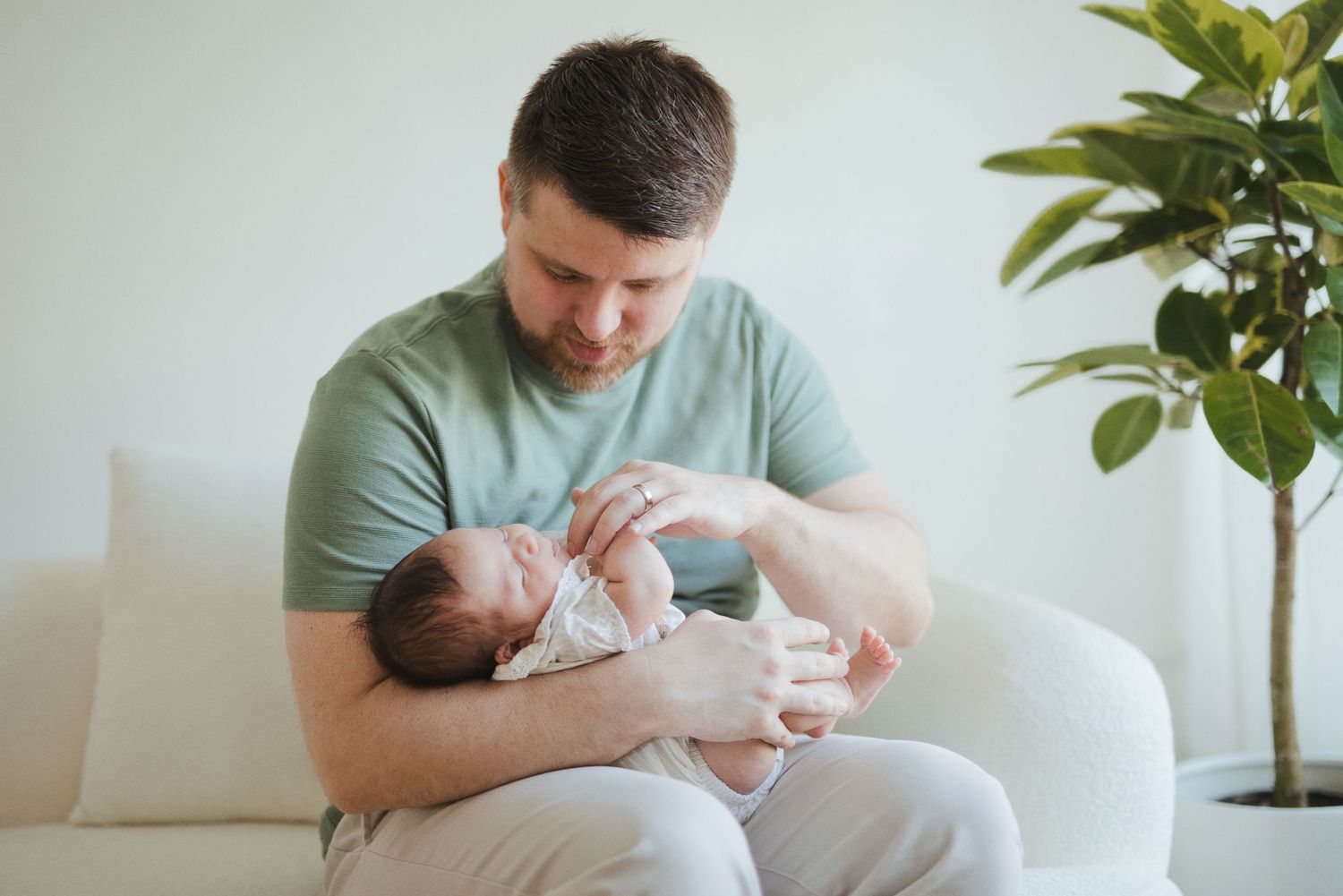 Father cradling newborn baby on white sofa at home, bonding with infant in bright modern living room.