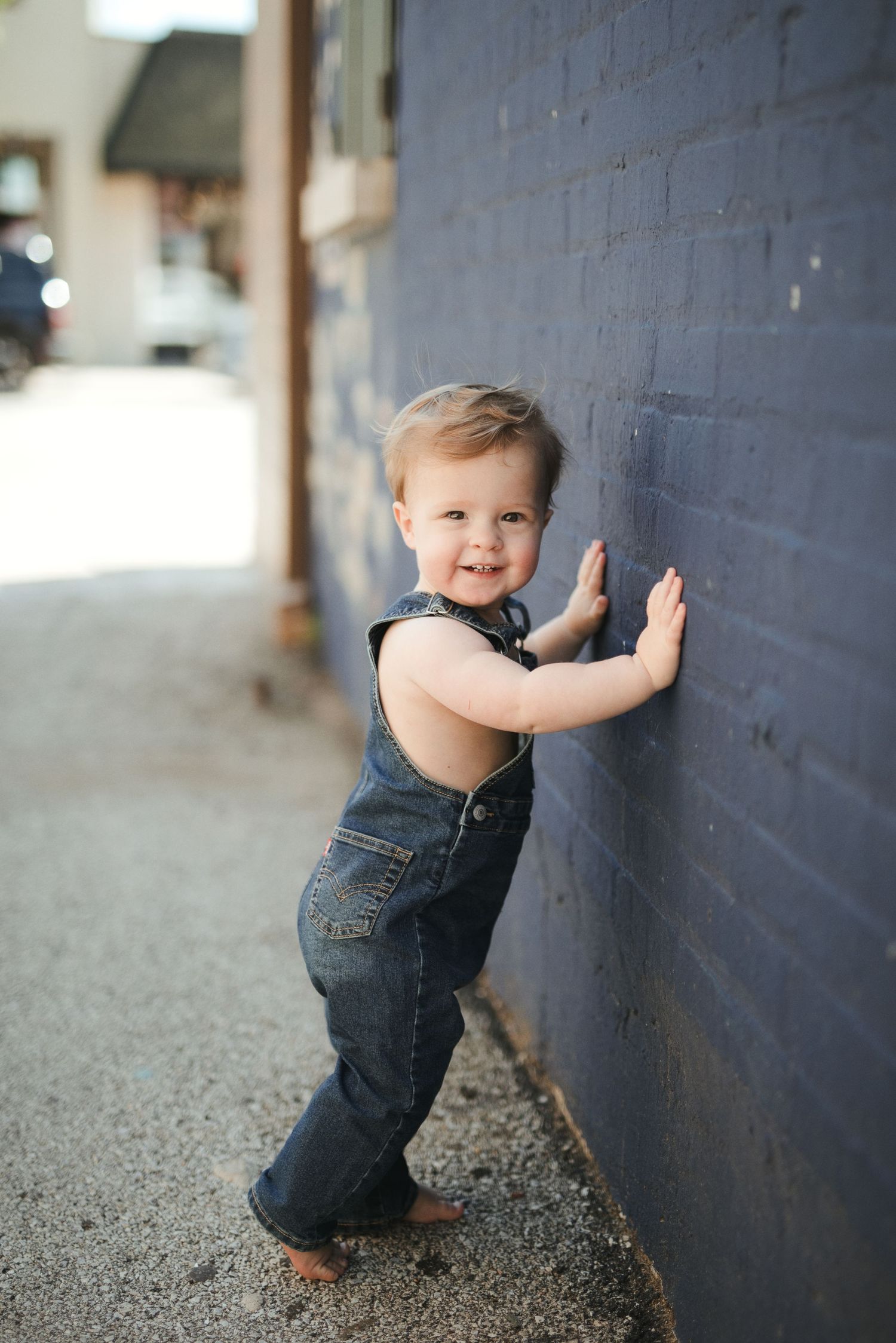 Smiling toddler in denim overalls leaning against a blue brick wall outdoors.
