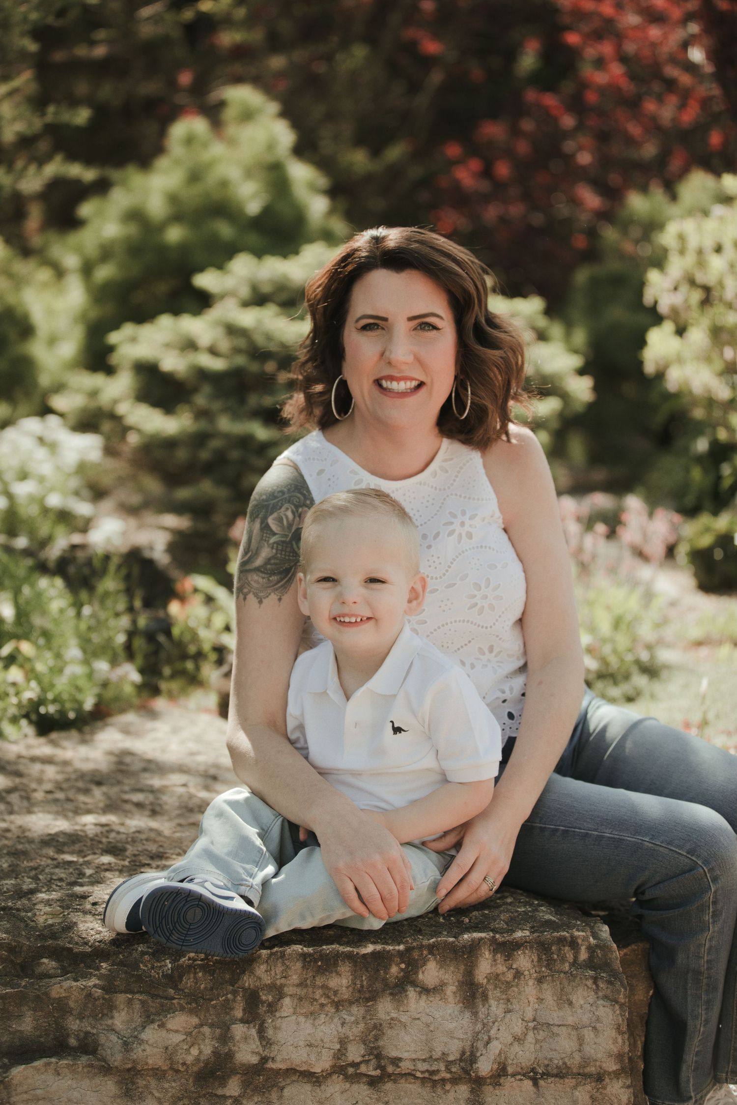 A smiling woman with dark hair and tattoos sits outdoors on stone steps with a young blonde toddler boy in a garden setting.