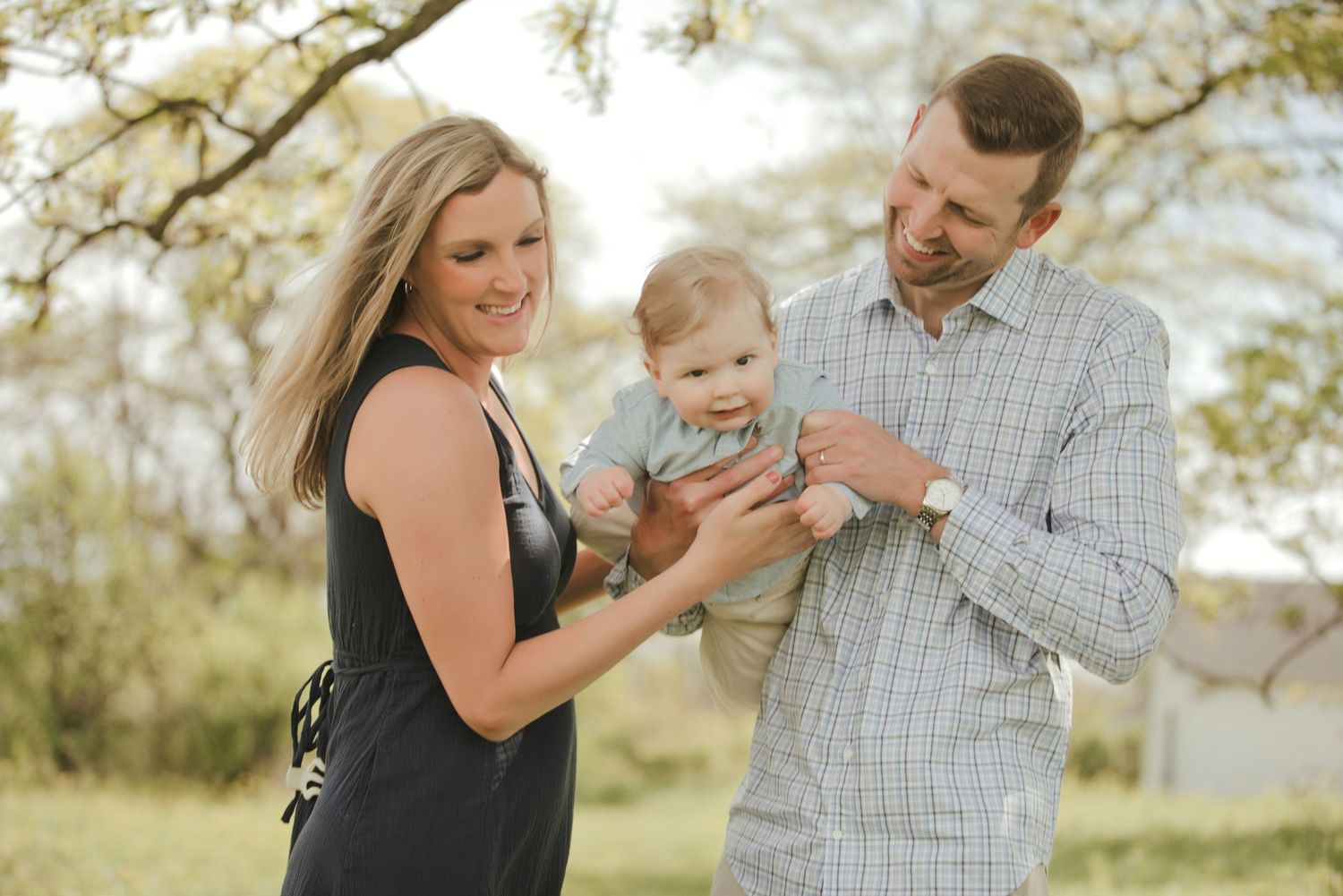 A happy young family with a baby outdoors under blooming trees in a warm, natural setting.