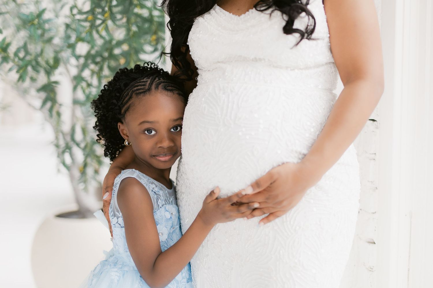 Young girl in blue dress hugging pregnant woman in white lace gown during maternity photo session.