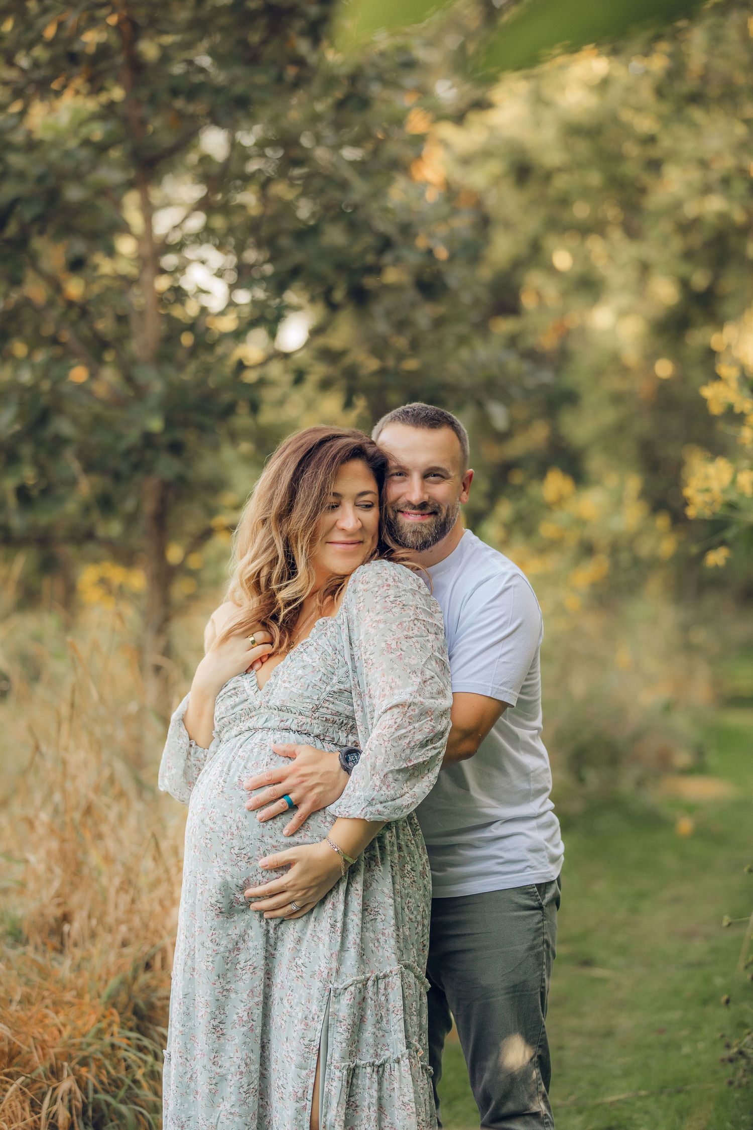 A couple embraces in a romantic outdoor maternity photo session surrounded by greenery.