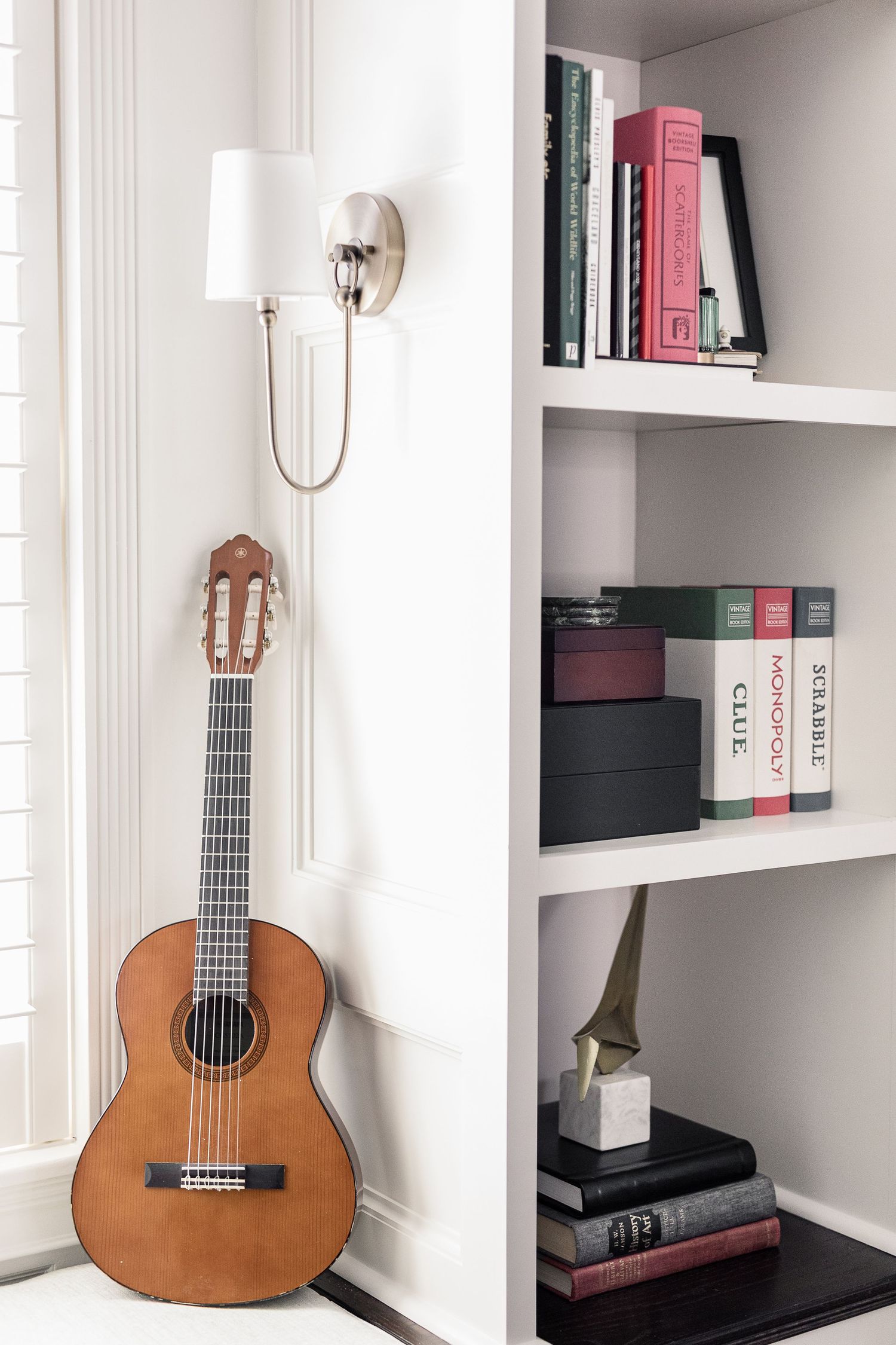 Classical guitar leans against wall beside built-in bookshelf with books and wall sconce light fixture.
