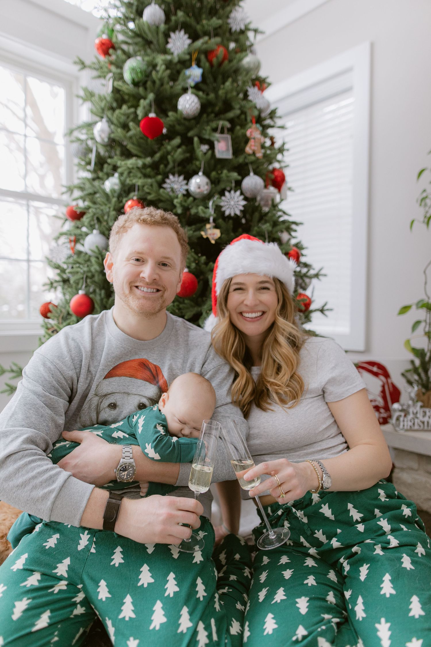 Family wearing matching green Christmas tree pajamas poses in front of decorated tree in bright white room.