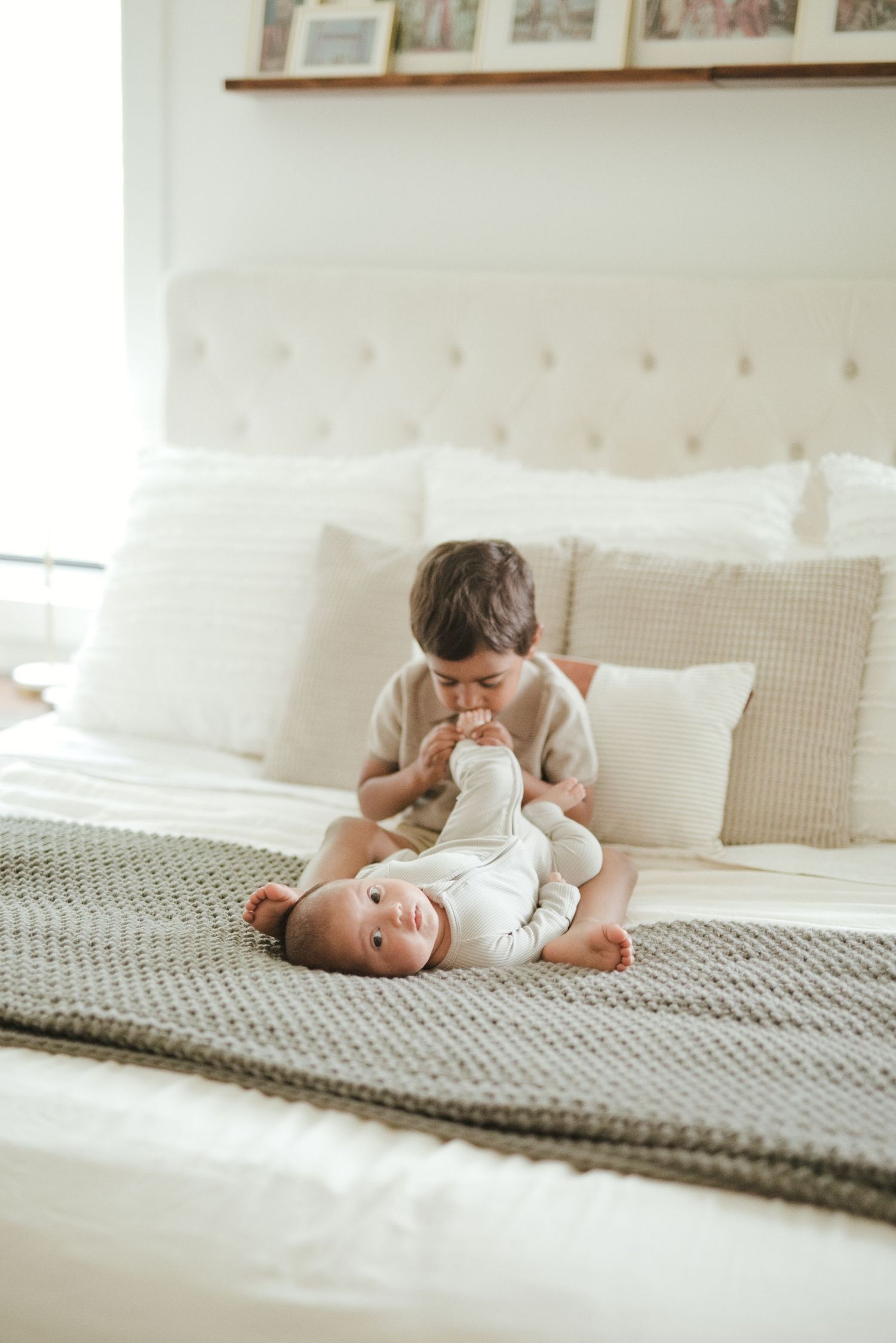 Two babies in white onesies playing together on a cozy white bed with soft pillows and a knit blanket.