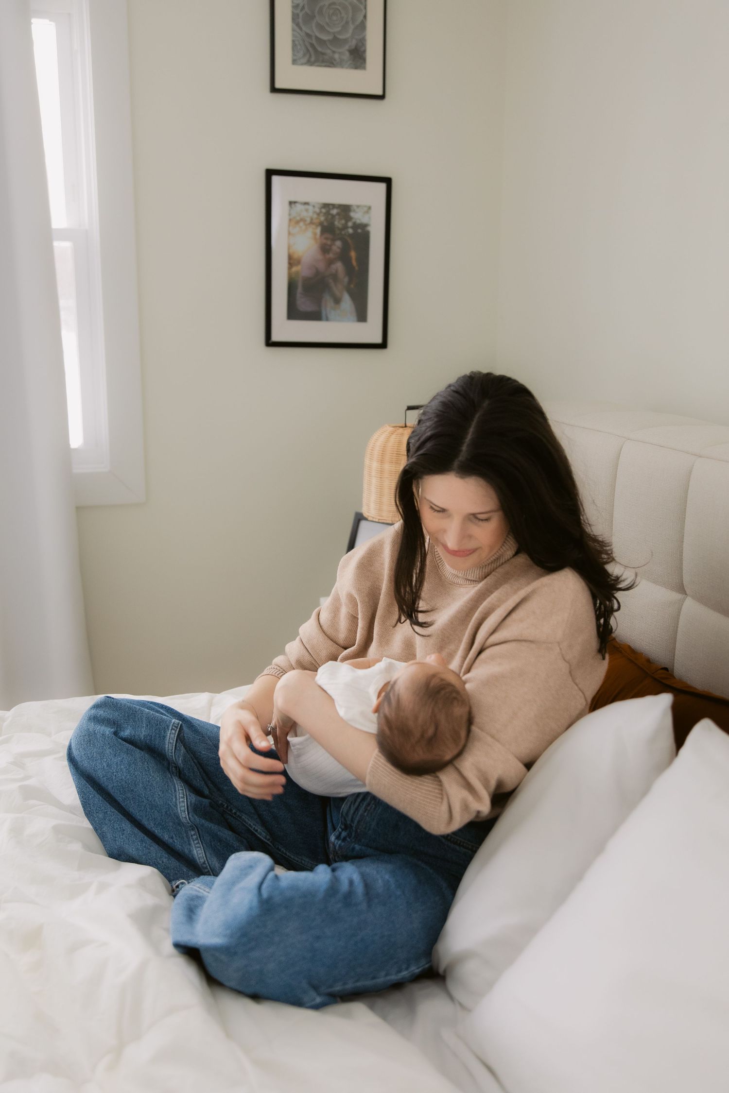 A parent in beige sweater cuddles with infant on white bedding while looking down lovingly.