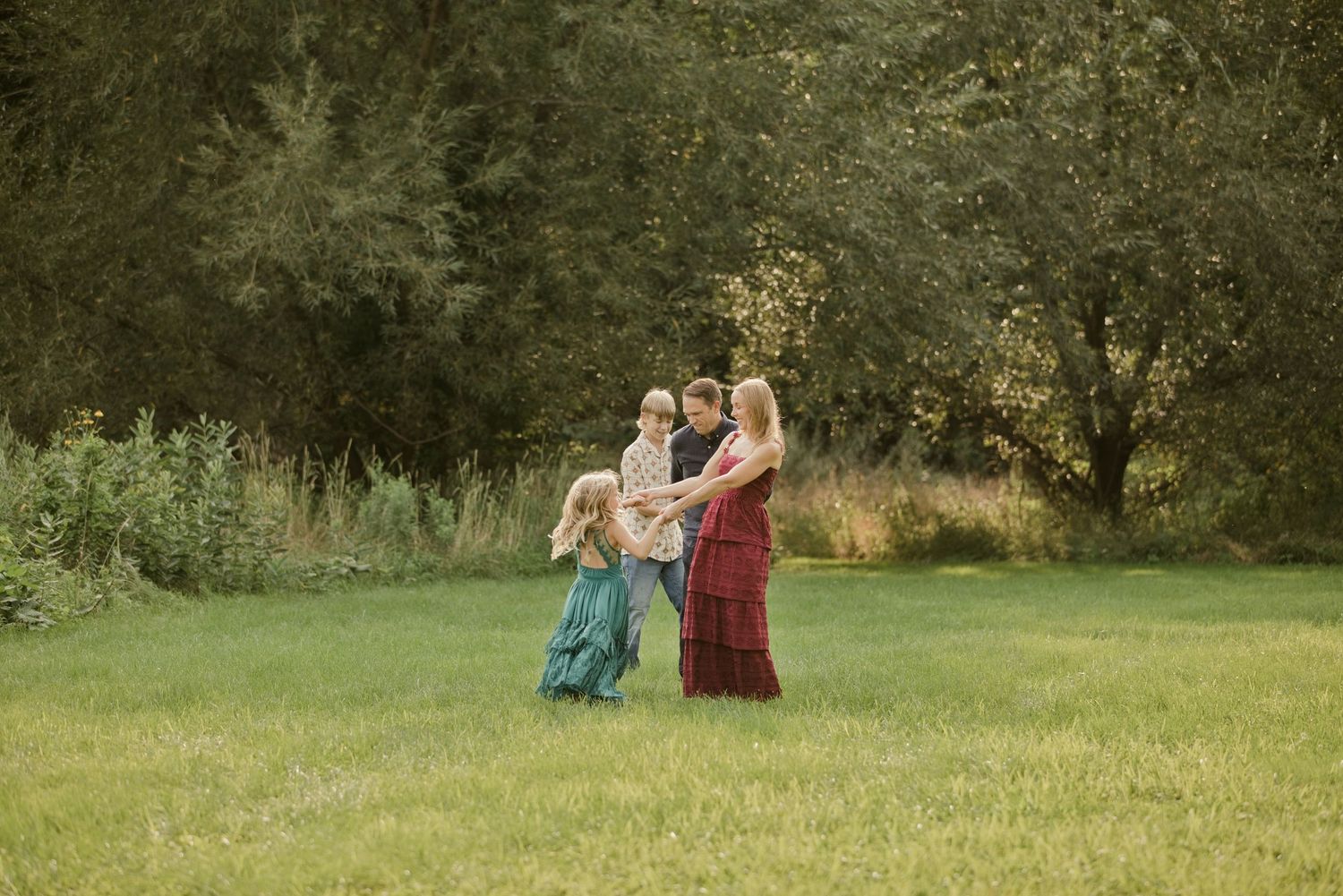 A mother in a red dress holds her baby while two young children play around her in a lush green meadow.