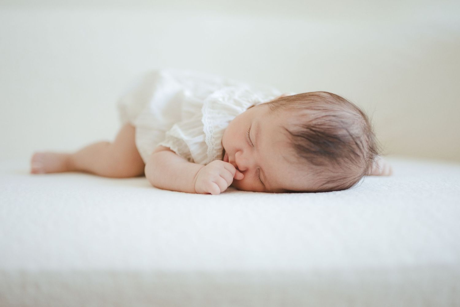 Peaceful newborn baby sleeping on white surface wearing a white lace outfit with dark hair visible.