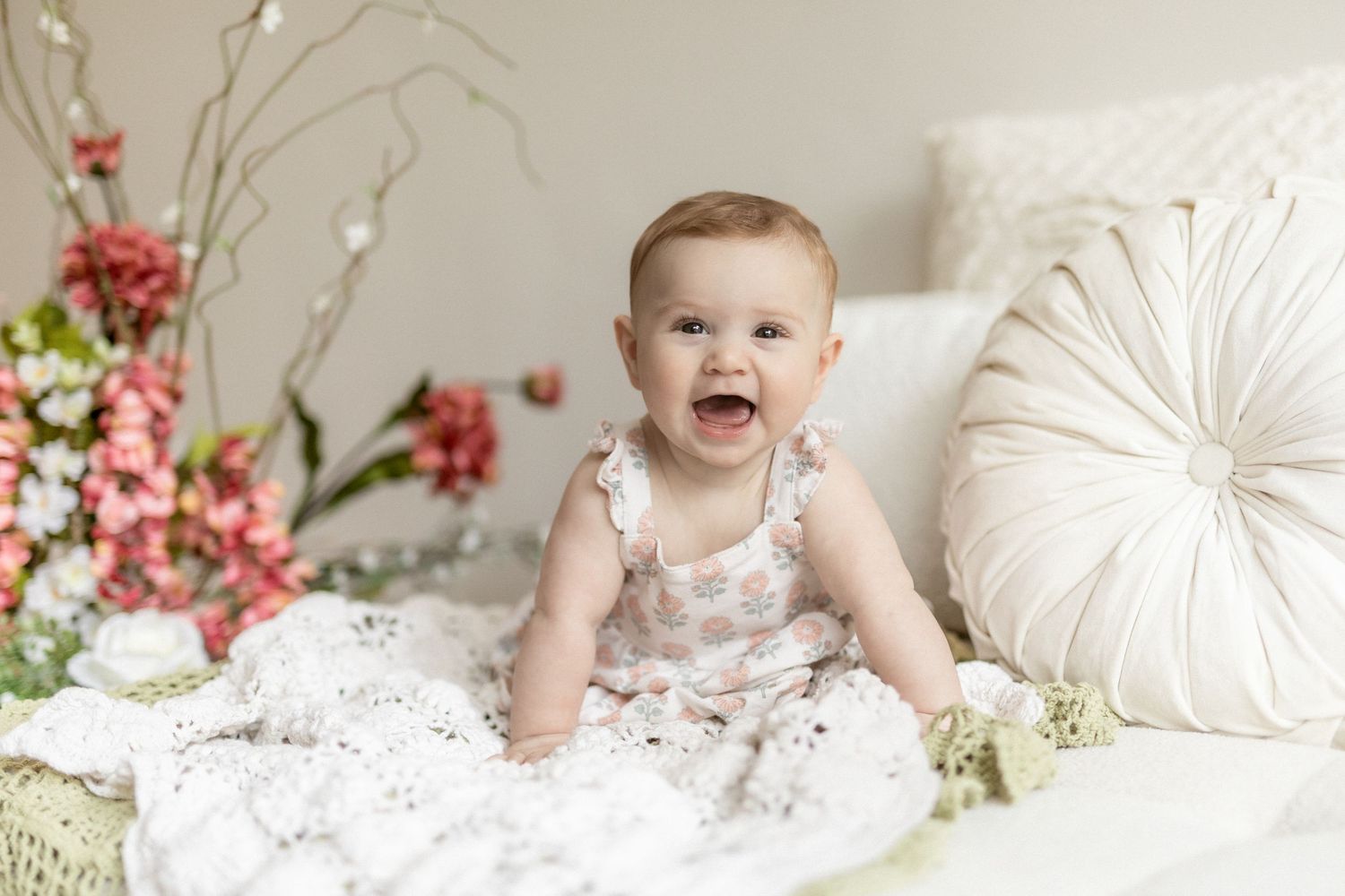 Happy baby girl in floral romper sitting on white lace blanket surrounded by pink flowers and soft pillows.
