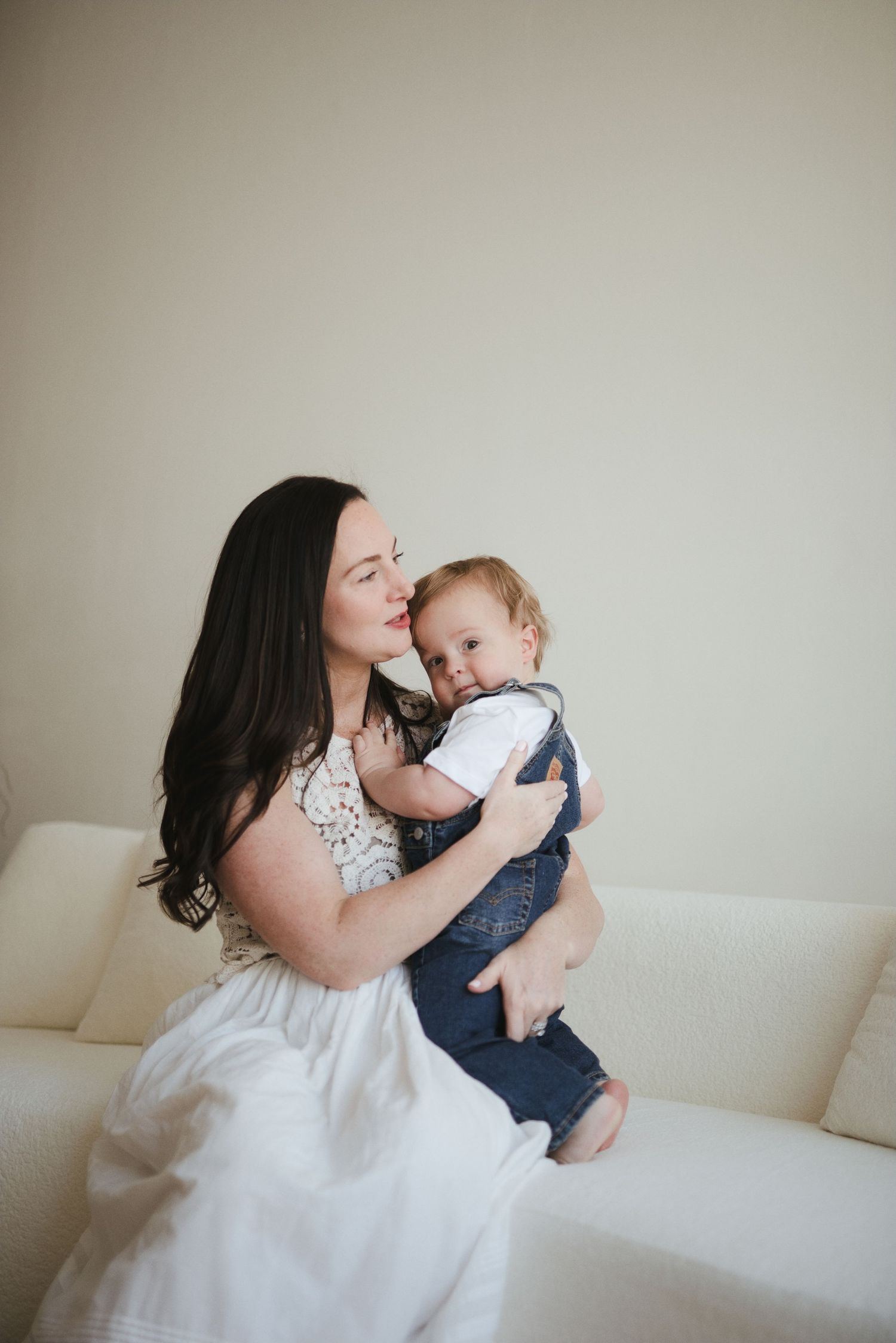 Dark-haired mother in white dress cuddling and kissing her smiling baby boy on a white couch.