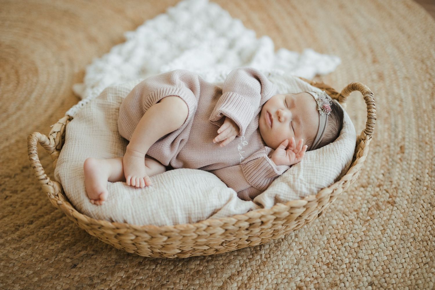 Newborn baby sleeping peacefully in a wicker basket with white blanket, wearing a pink knit outfit and headband.