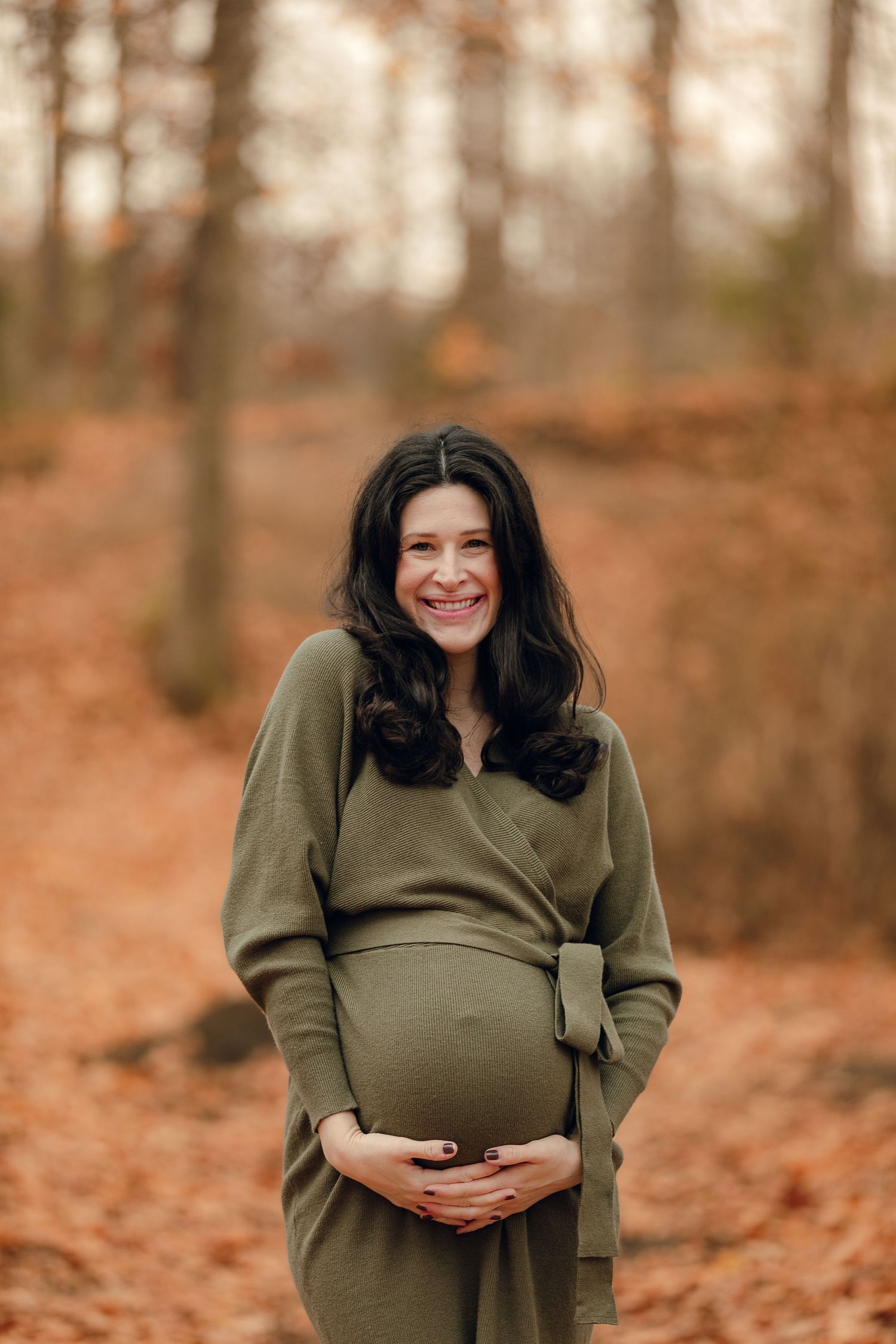 Expectant mother in olive green dress poses during fall maternity session surrounded by autumn leaves.