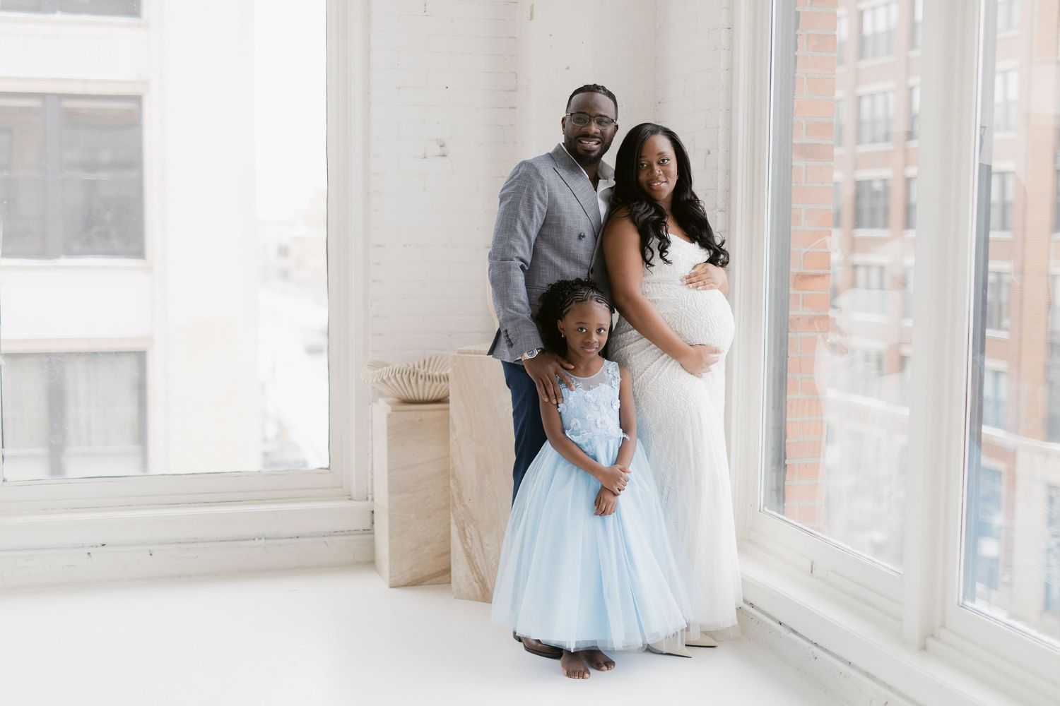 A pregnant woman in a white gown poses with her husband and young daughter in a bright studio setting.