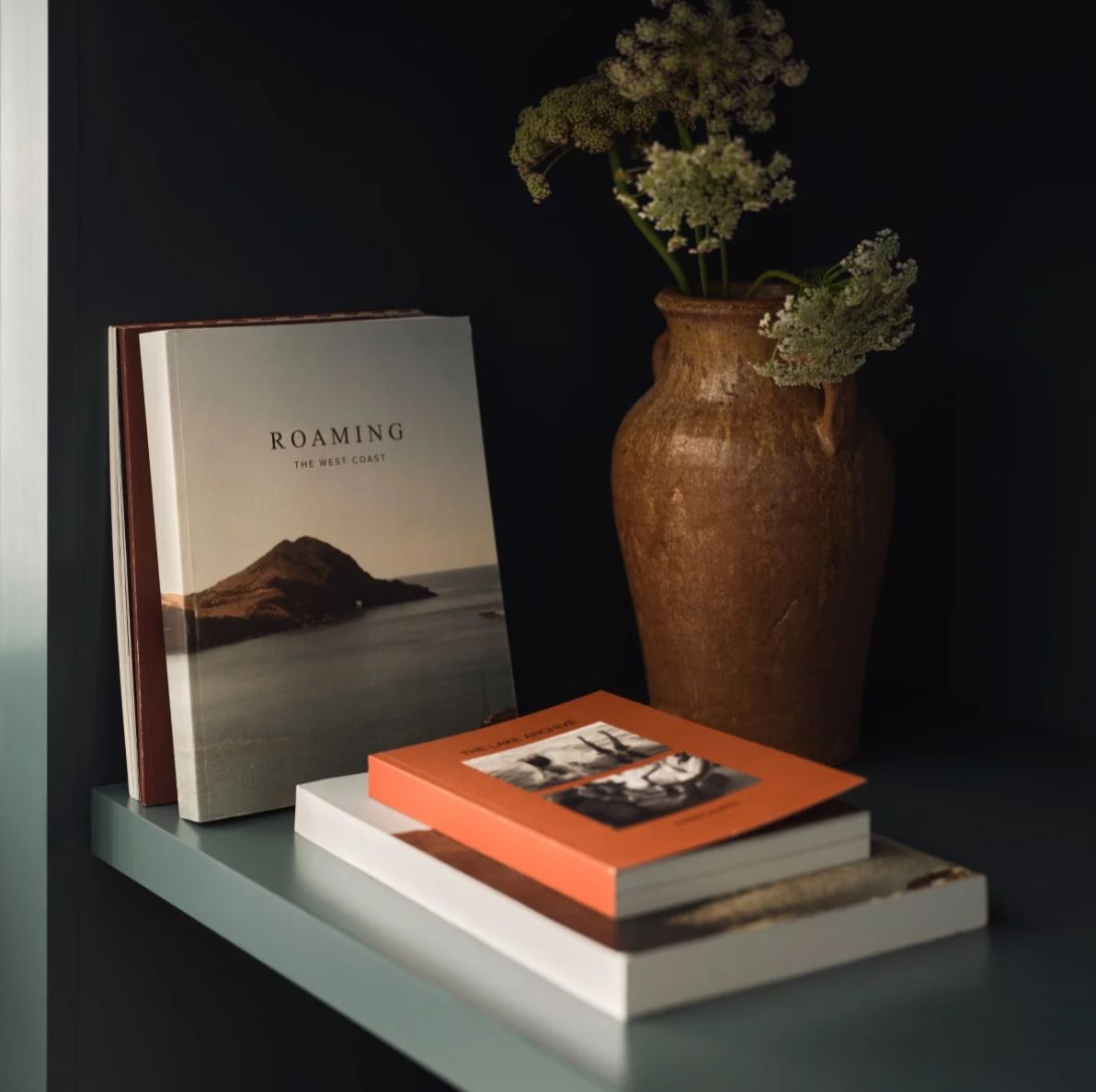 Stack of decorative books including Roaming beside a rustic ceramic vase with dried flowers on a shelf.