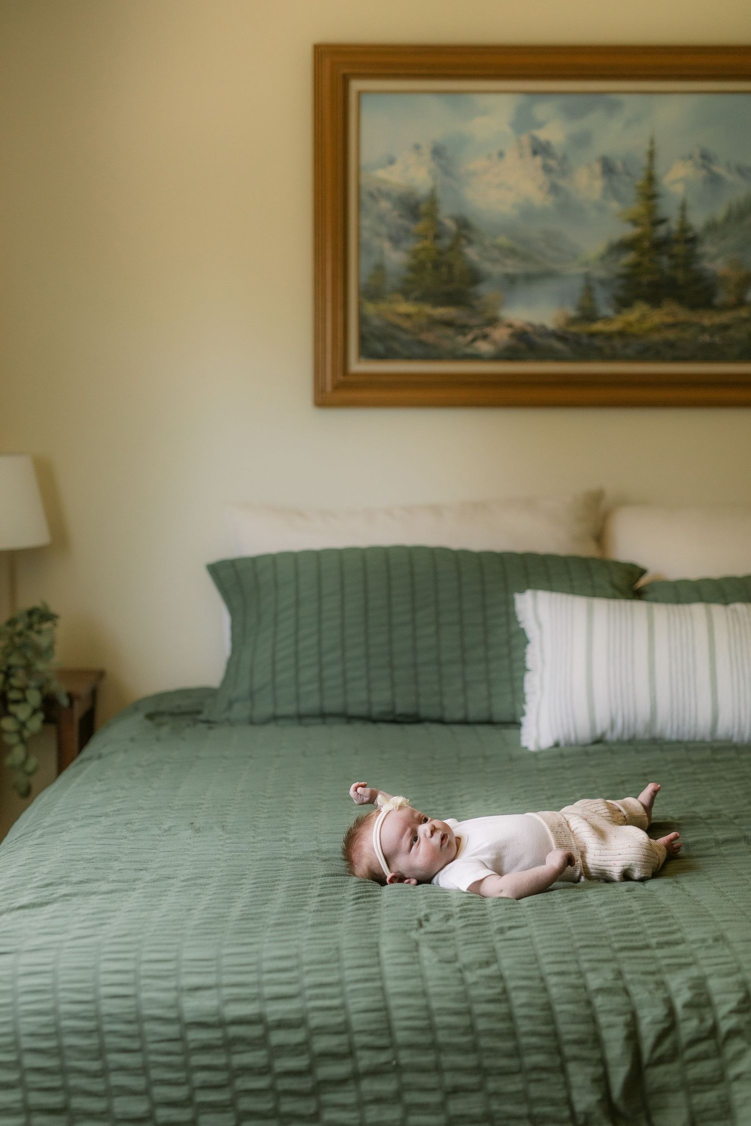 A sleeping infant rests on a sage green quilted bedspread with landscape artwork on the wall.
