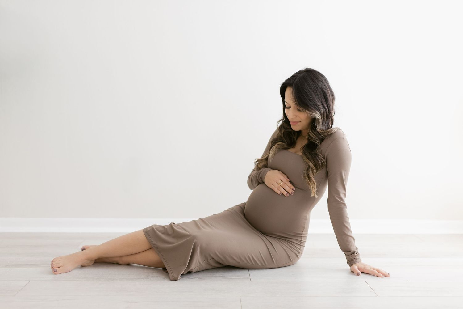 A pregnant woman in a brown dress sits on a white floor while looking down at her belly in a minimalist maternity setting.