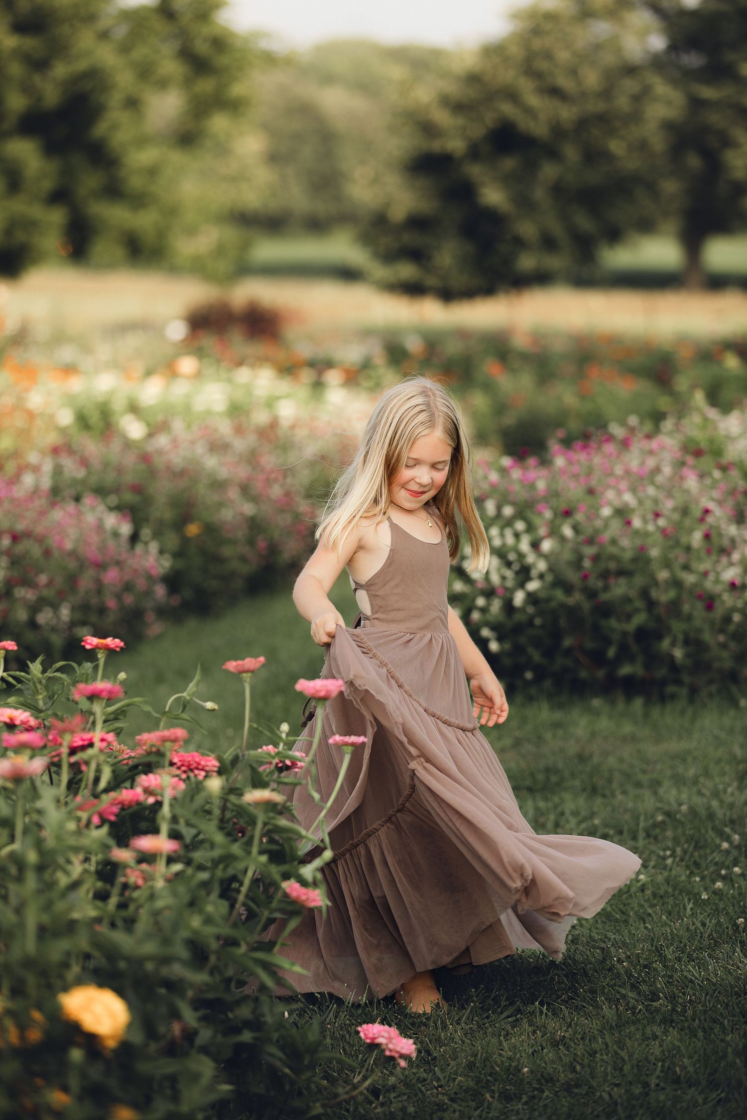 Young child twirling in mauve dress among colorful garden flowers at sunset.
