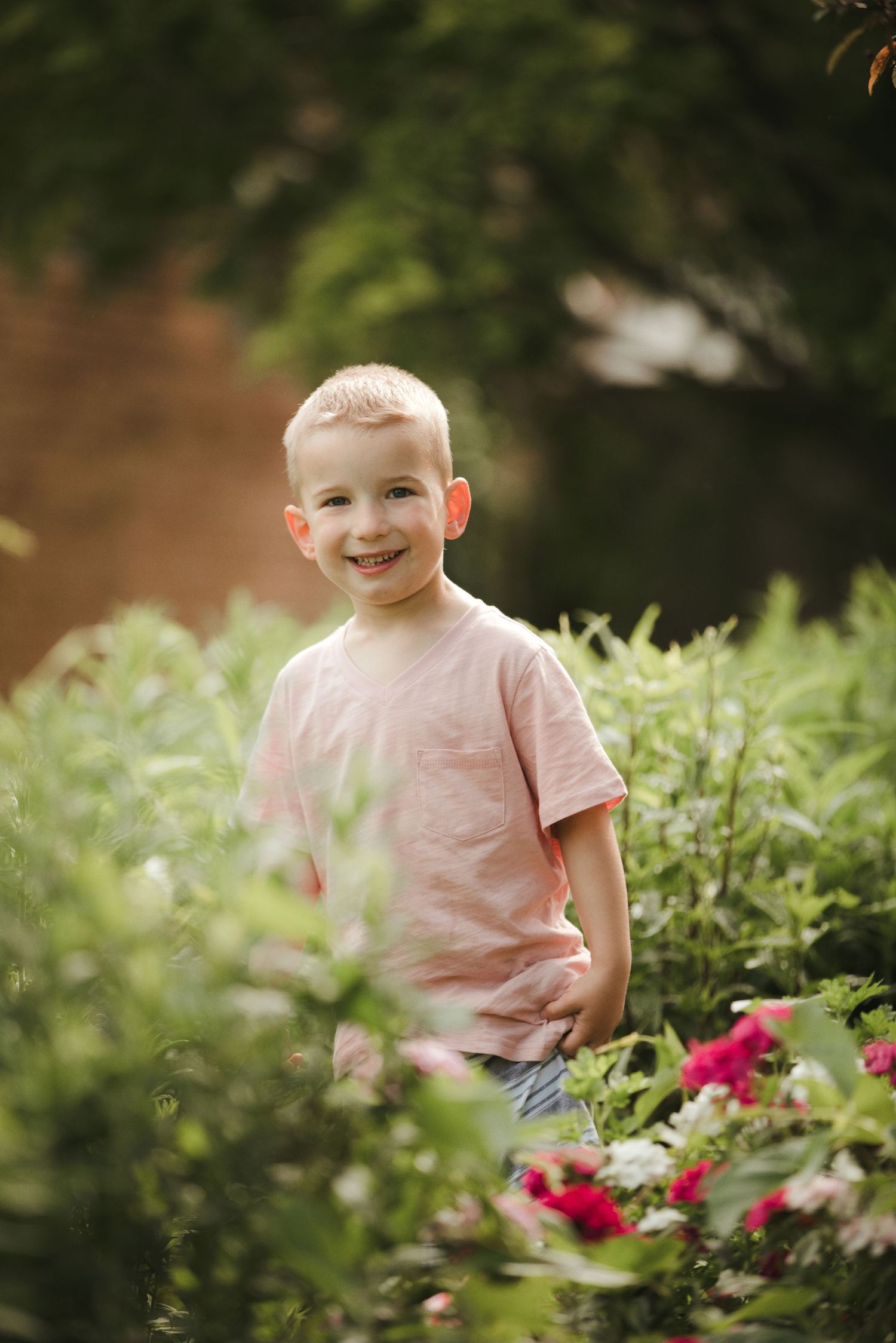 Smiling young boy in pink t-shirt standing among colorful garden flowers and lush green plants outdoors.