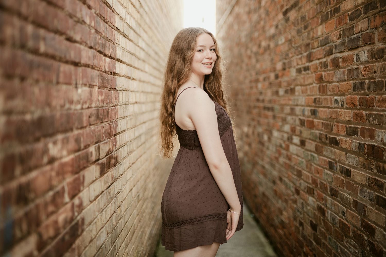 Young woman in brown dress smiling over shoulder in brick alley, long auburn hair, senior portrait photography.