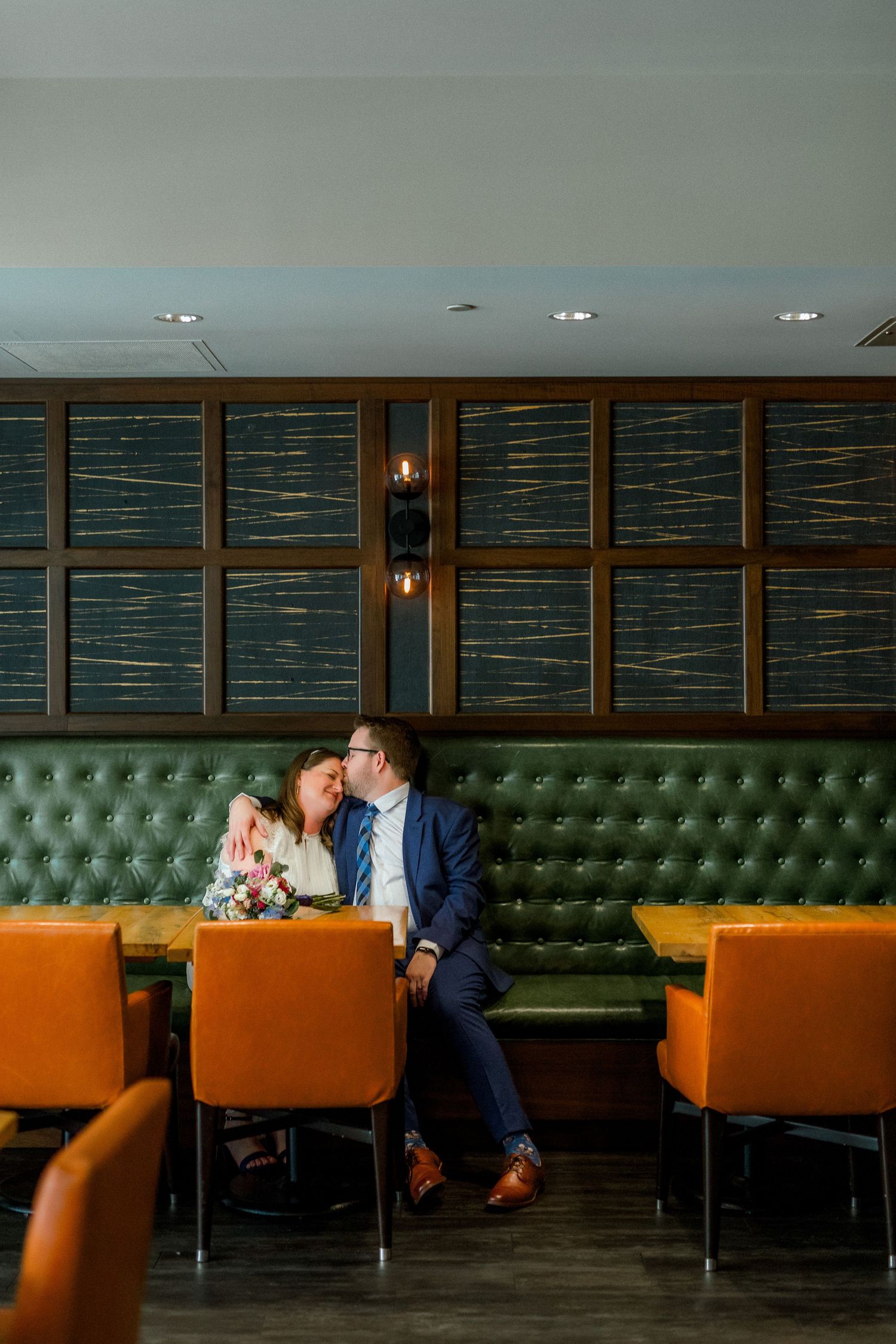 Couple sharing a romantic moment on a tufted green booth in an upscale restaurant with orange chairs.