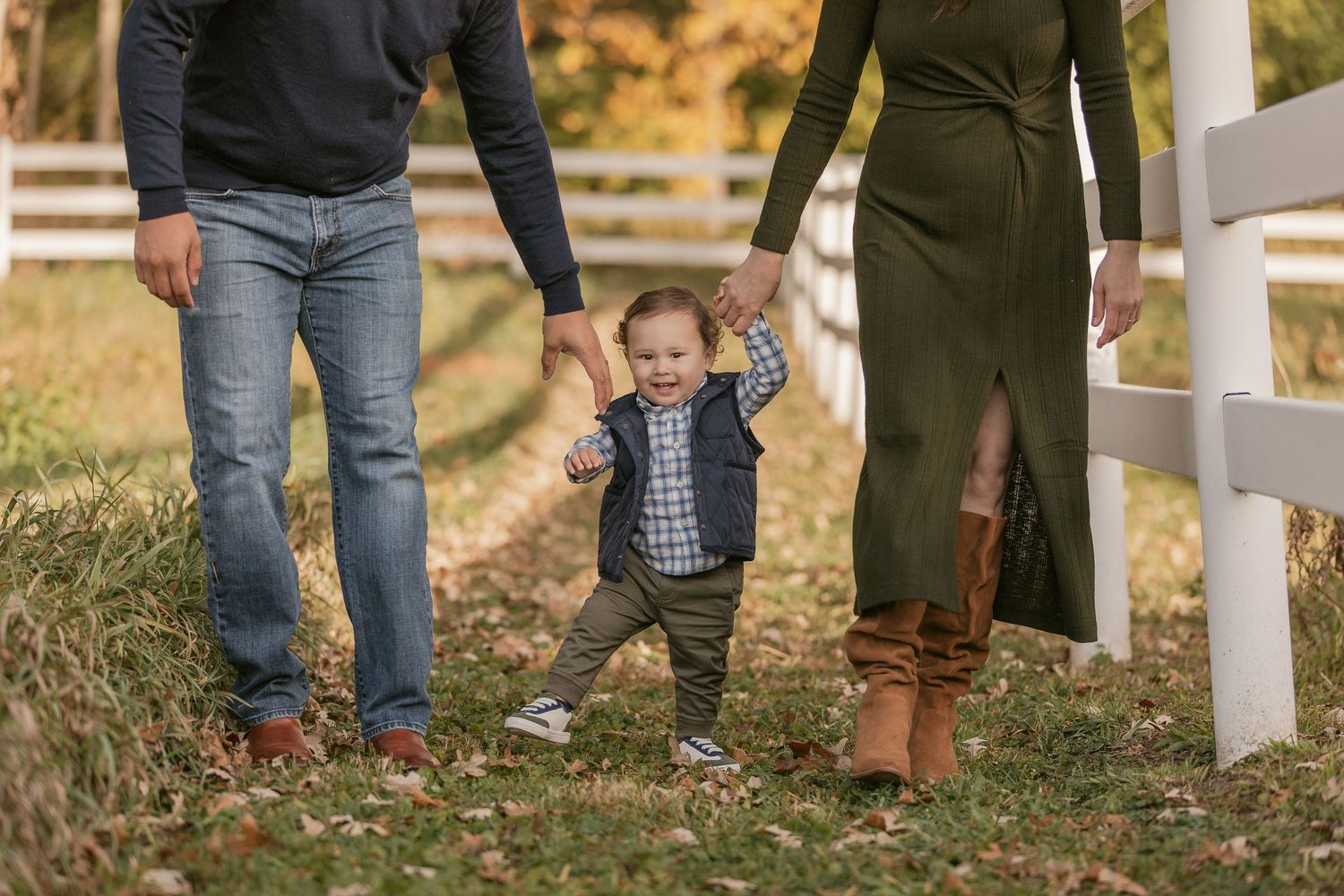 A toddler walks between parents holding hands outdoors near a white fence during a fall family photo session.