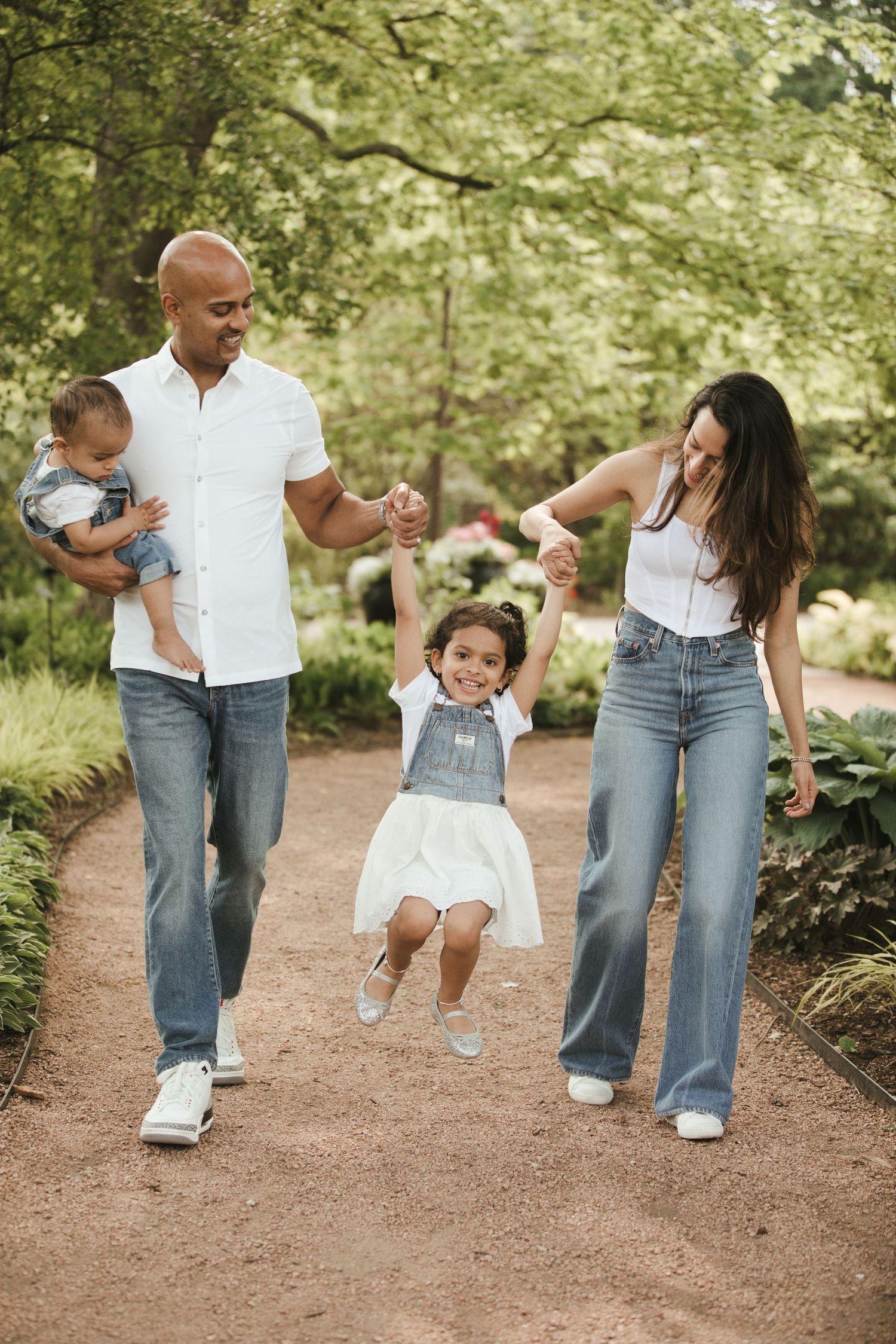 A joyful family walks a garden path, parents swinging their daughter while dad holds a baby, all in casual denim outfits.
