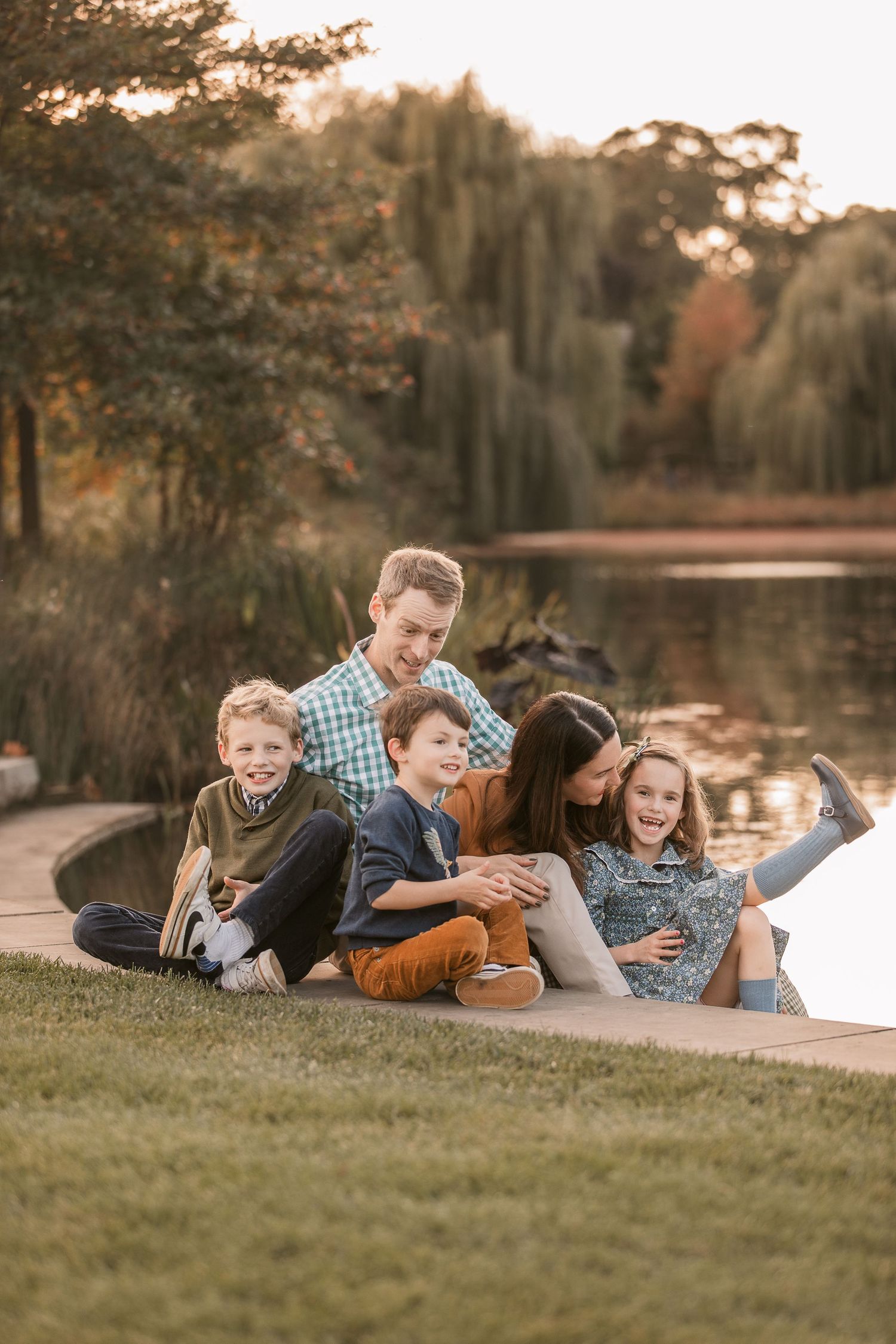 Happy family of five sitting together on grass by a pond, laughing and embracing during a fall outdoor photo session.