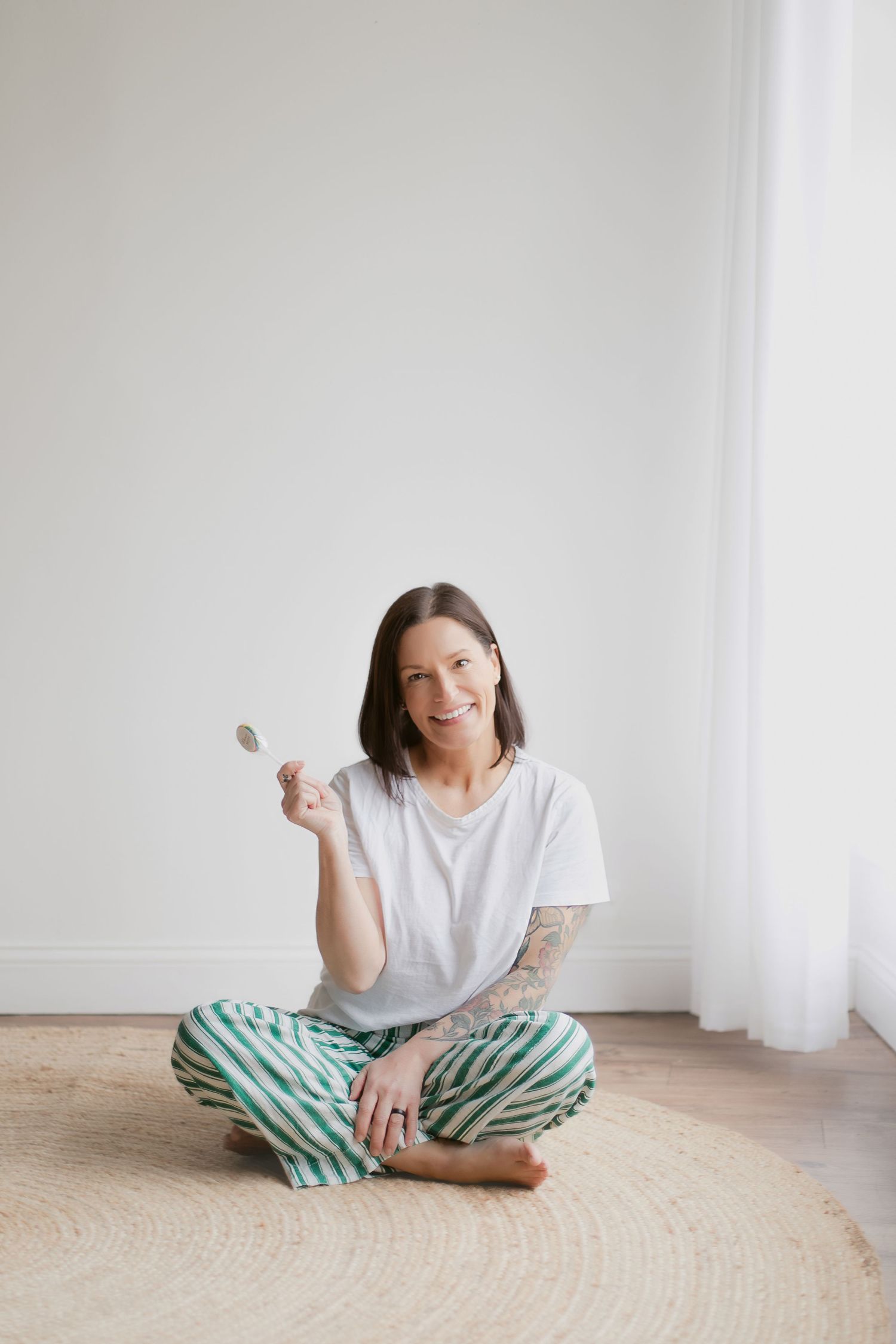 Smiling woman with tattoos sitting cross-legged on a round rug, wearing white tee and striped pants, holding a wooden spoon.