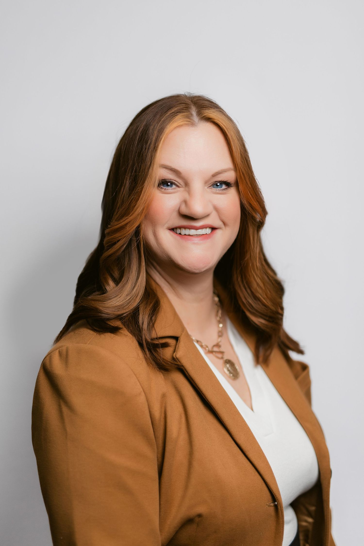 Professional headshot of person in brown blazer and white blouse against grey background.