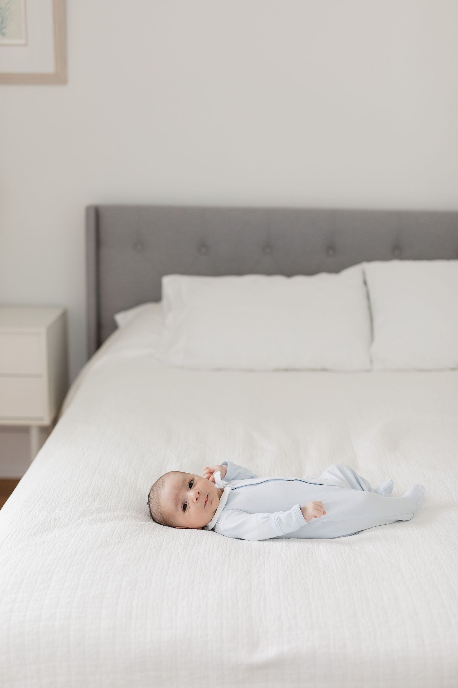 Baby in light blue outfit lying on white bedding in modern bedroom.