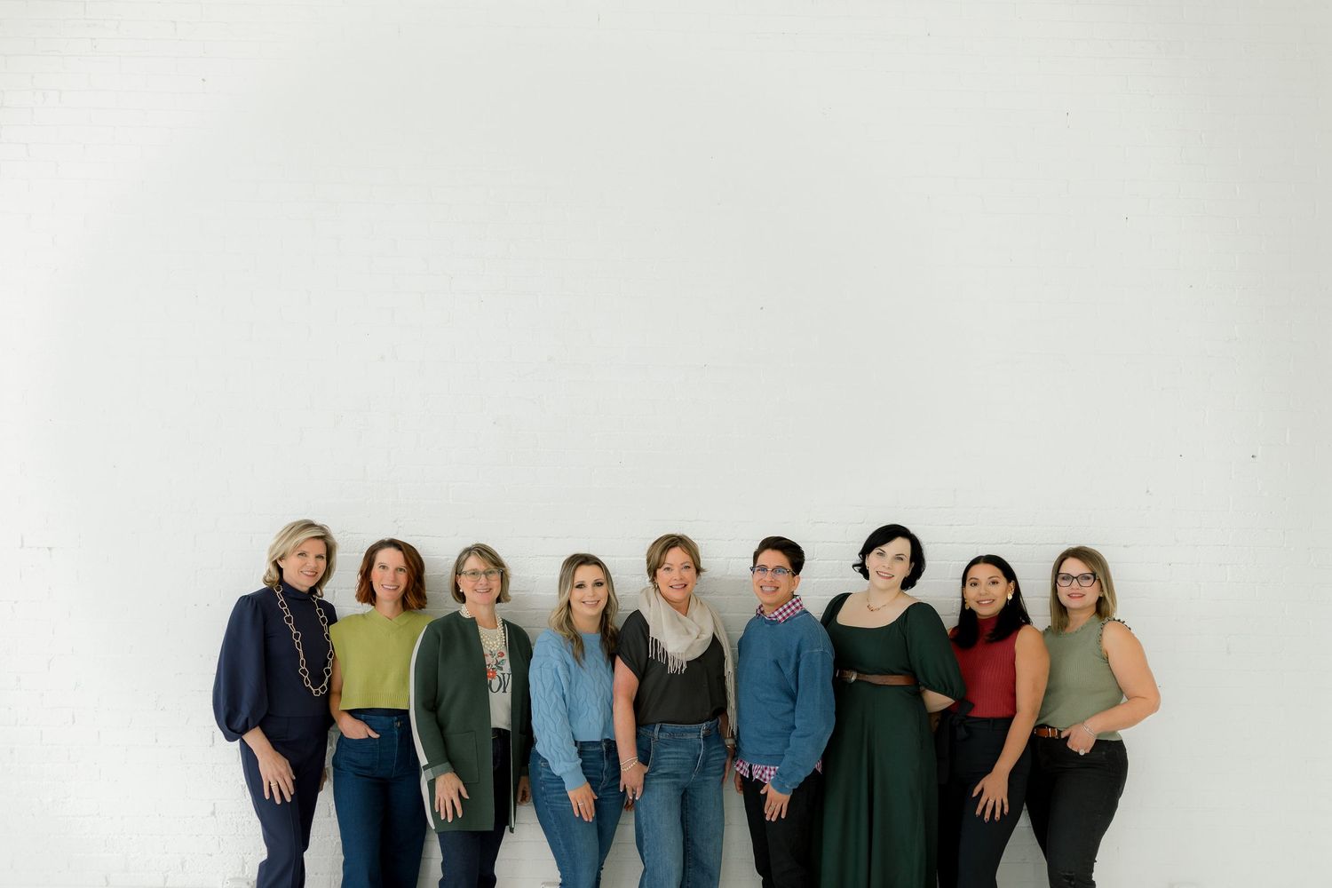 A group of nine people stand together against a white wall wearing casual clothing in earth tones and jewel colors.
