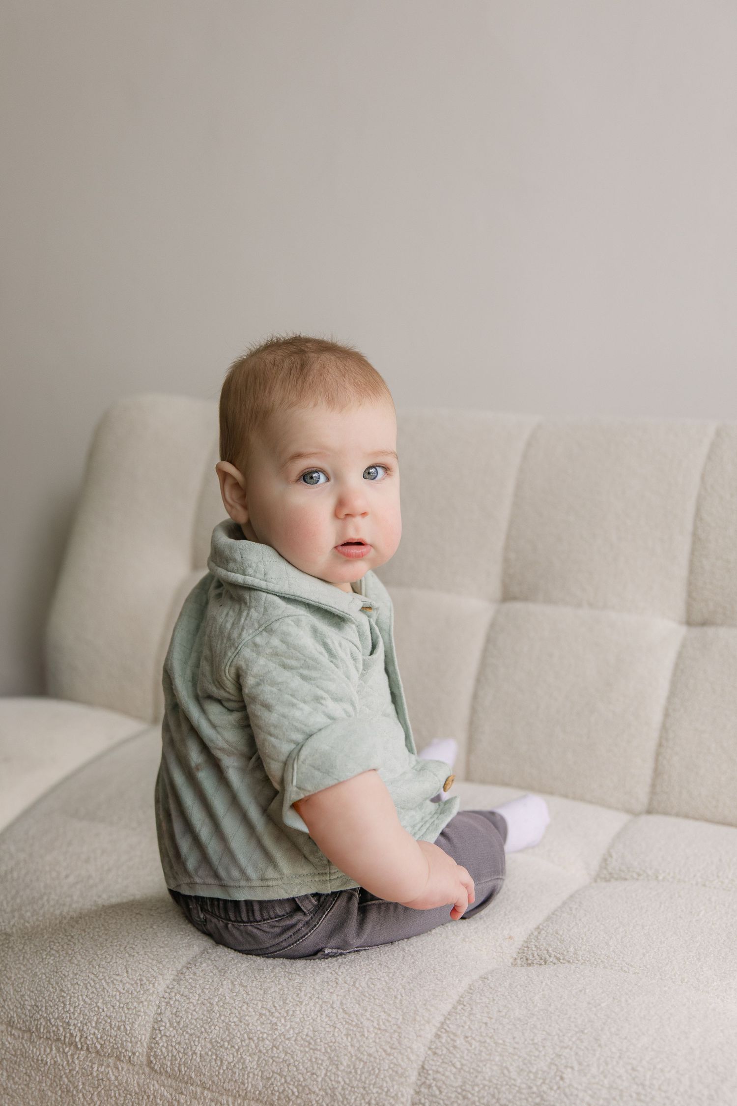 A baby in a mint green shirt sits on a light gray couch in a series of progressive poses.