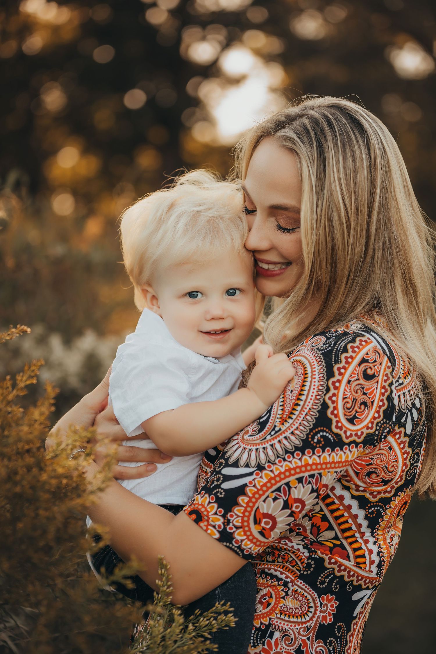 A tender moment captured in warm golden light shows a parent embracing a small child wearing white against an autumn backdrop.