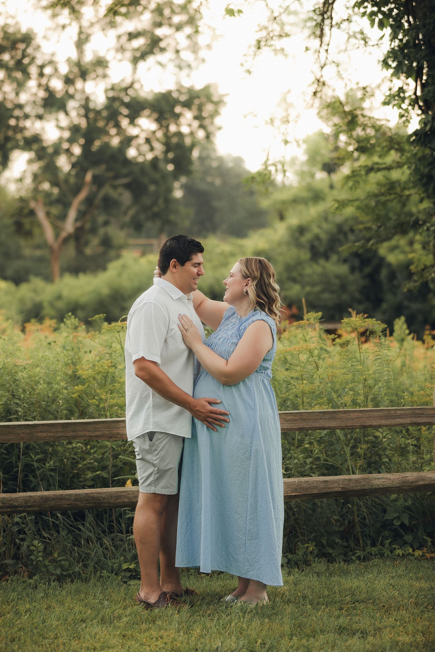 Couple sharing a tender moment outdoors, woman in blue maternity dress, man in white shirt, lush green background.