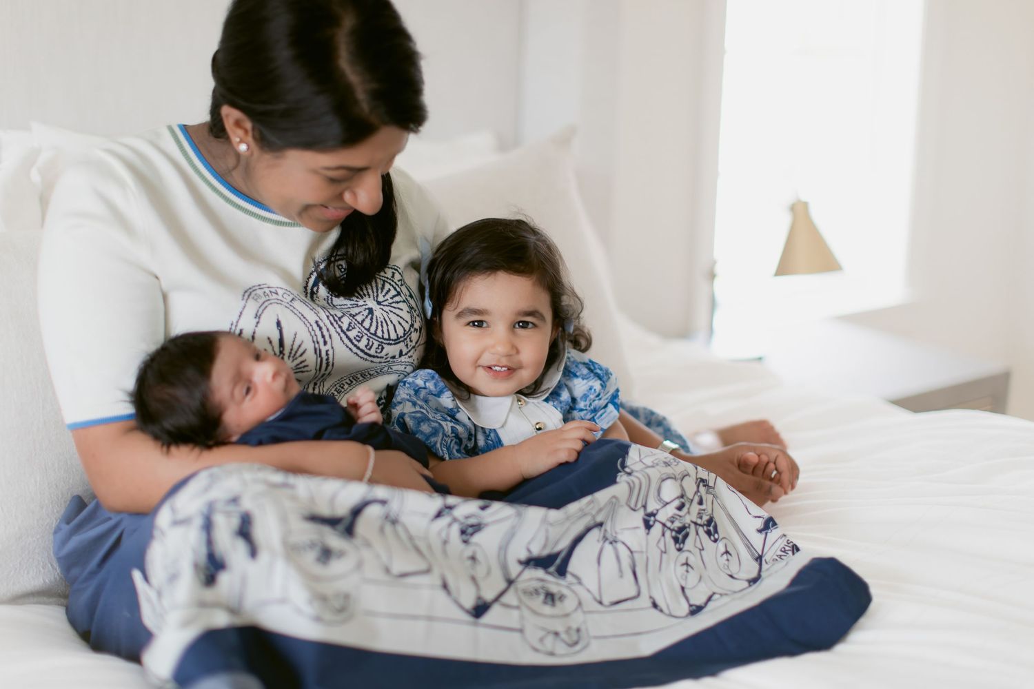 Mother sitting on bed with toddler daughter and newborn baby wrapped in a blue blanket.