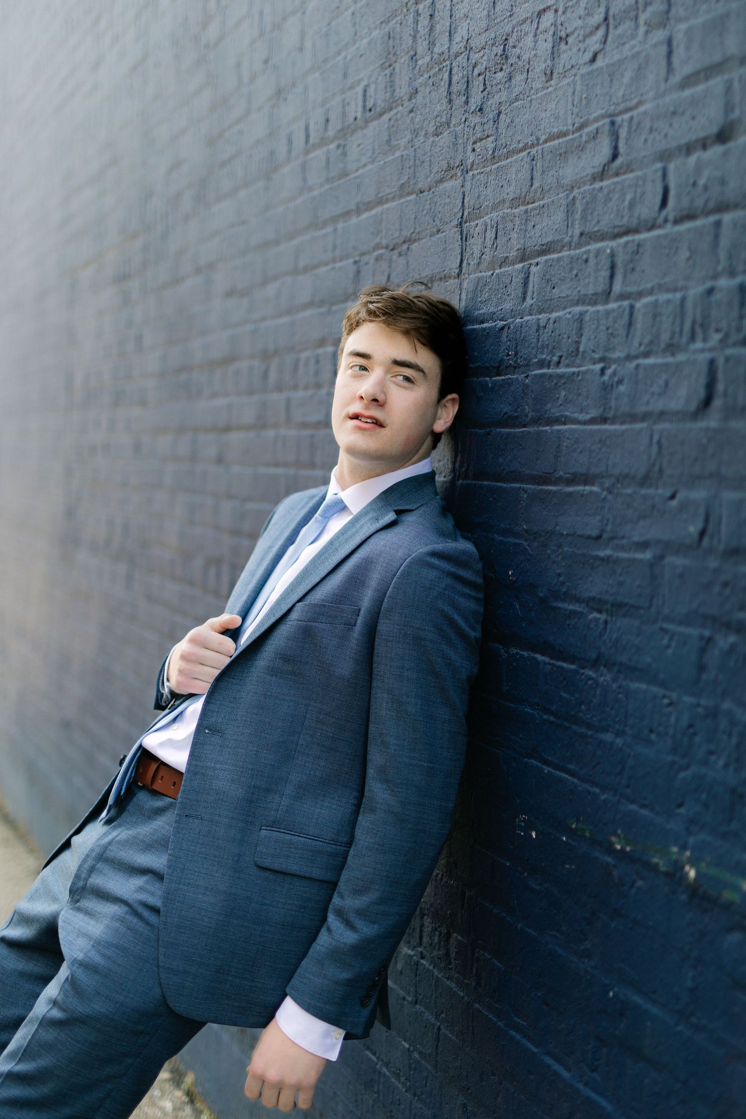 Young man in blue suit leaning against dark brick wall, posing confidently for a formal portrait photo.