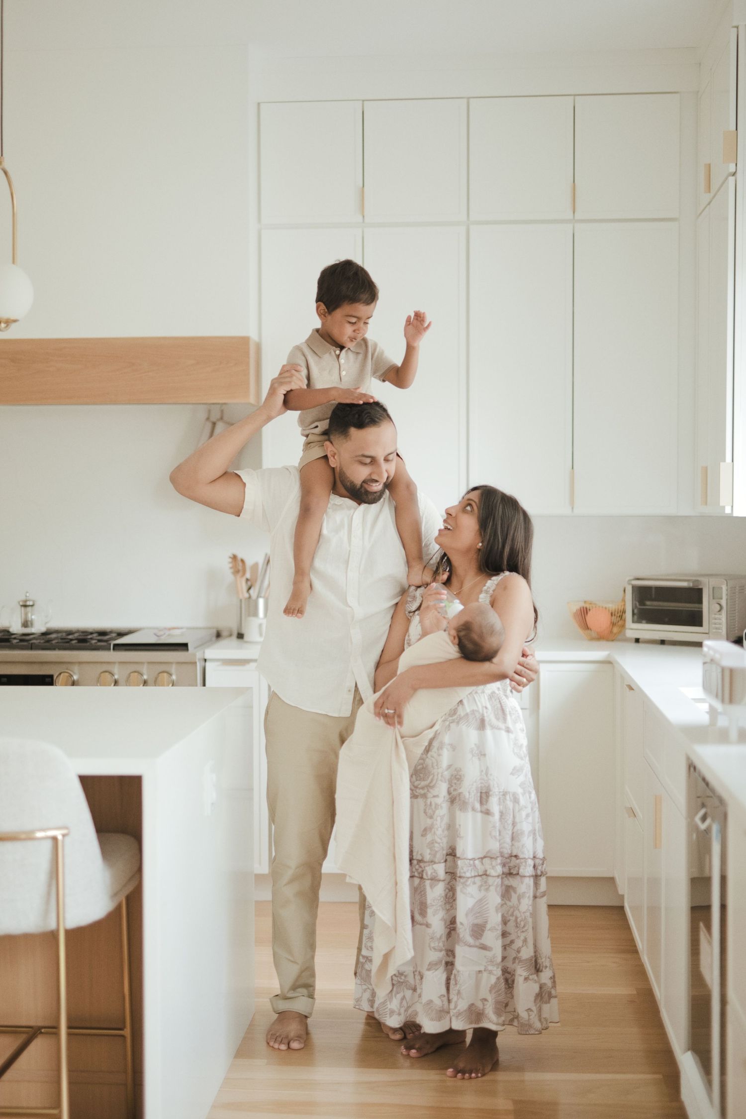 A happy family in a modern white kitchen with father carrying son on shoulders and mother holding newborn.