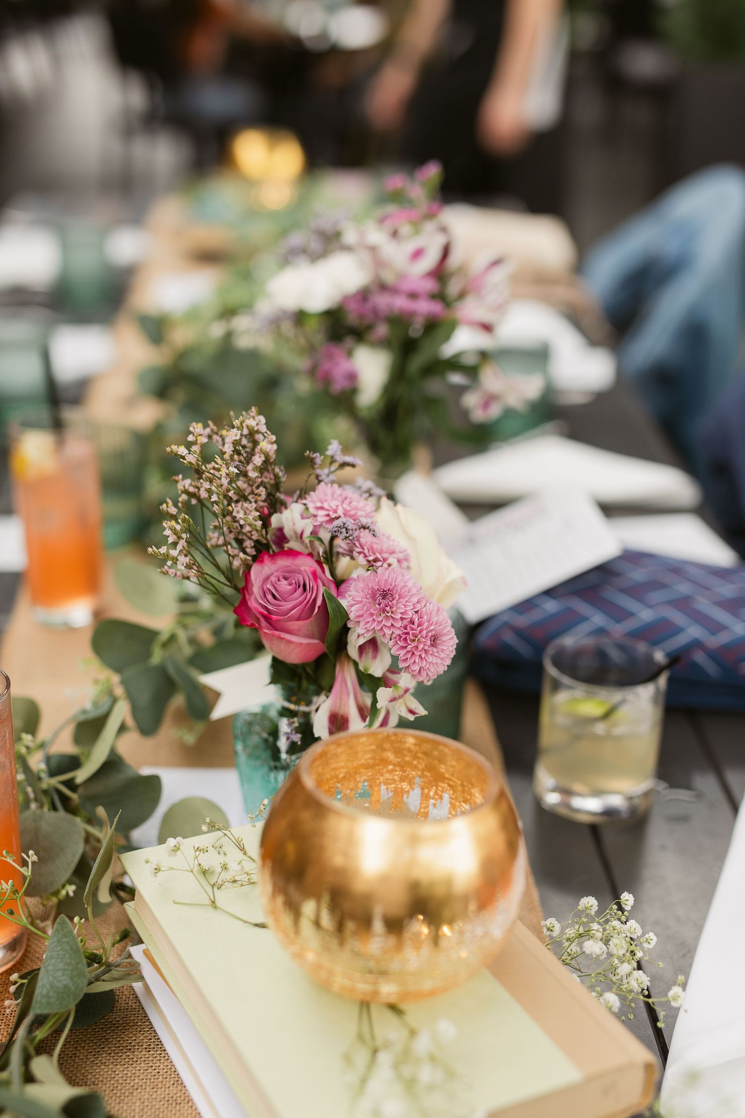 Elegant table setting with pink floral centerpieces, gold candle holder, stacked books, and drinks on a rustic wooden table.