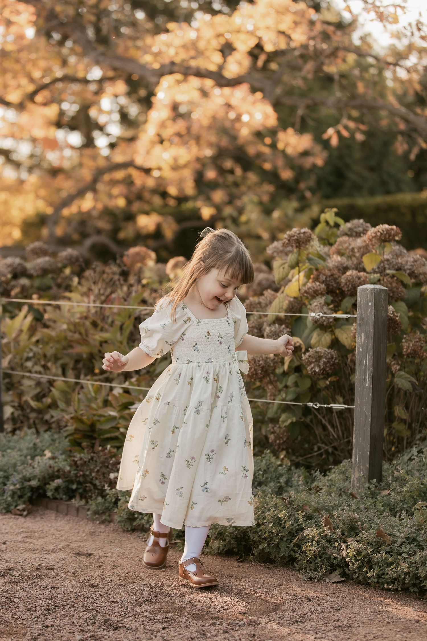 Small child in white dress exploring garden path during golden hour photo session.