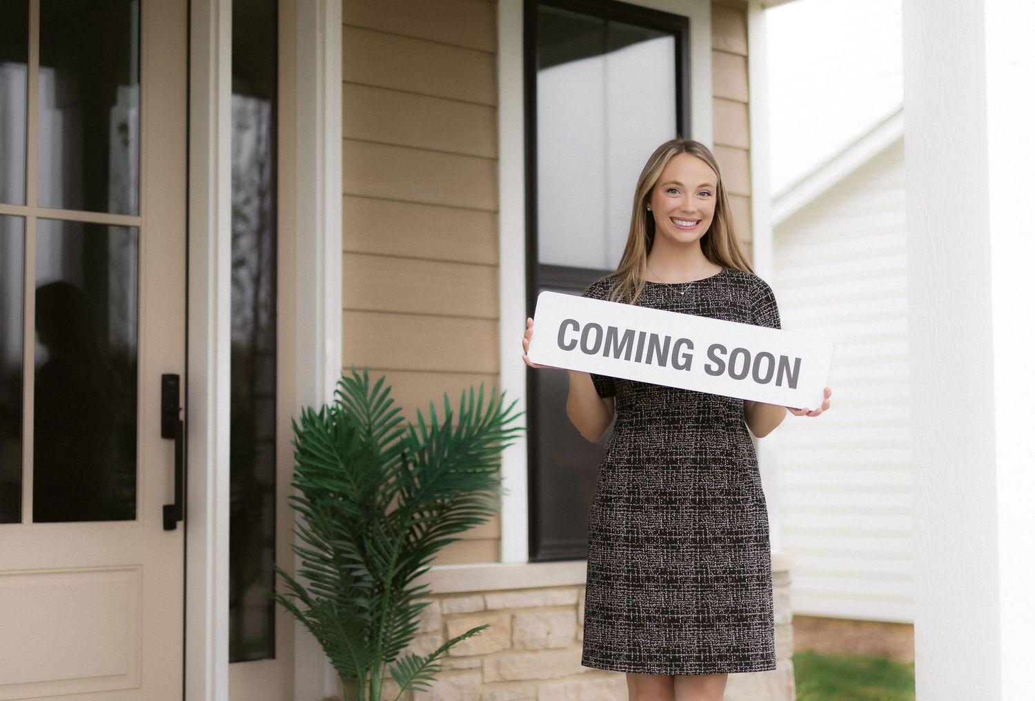 Professional stands in front of a beige house holding a 'Coming Soon' sign near a decorative palm plant.