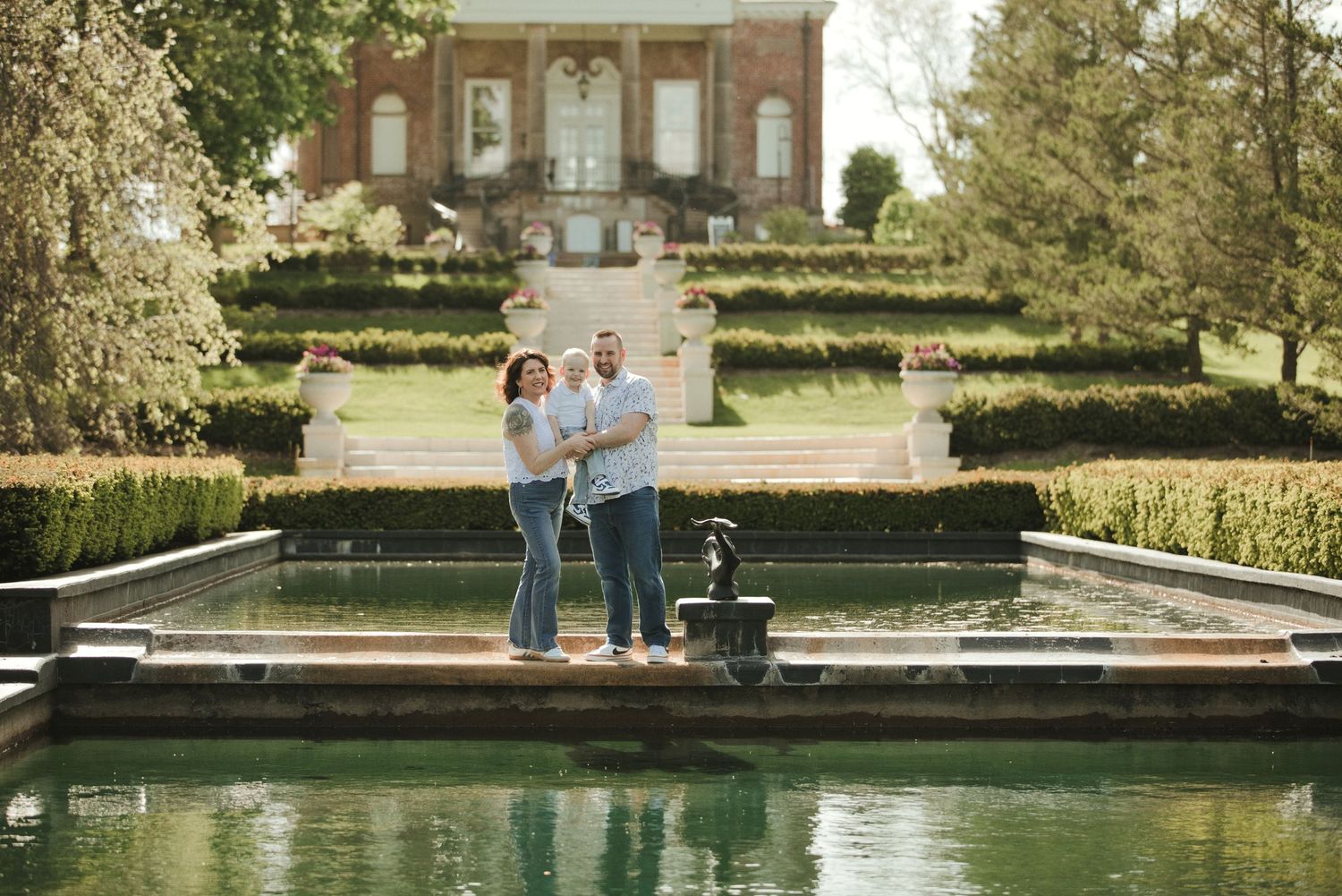 A couple stands together by a reflective pool in front of an elegant brick estate garden.